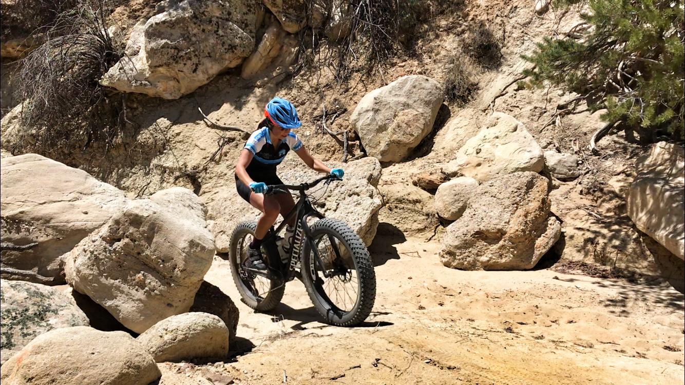 A young person riding a fat-tire mountain bike on a rocky, sunlit trail, navigating through large boulders and dry terrain. The cyclist is wearing a blue helmet and gloves, along with cycling attire, demonstrating skill and focus as they tackle a challenging path.  Double Slot Fat Bike Trail mountain bike trail.