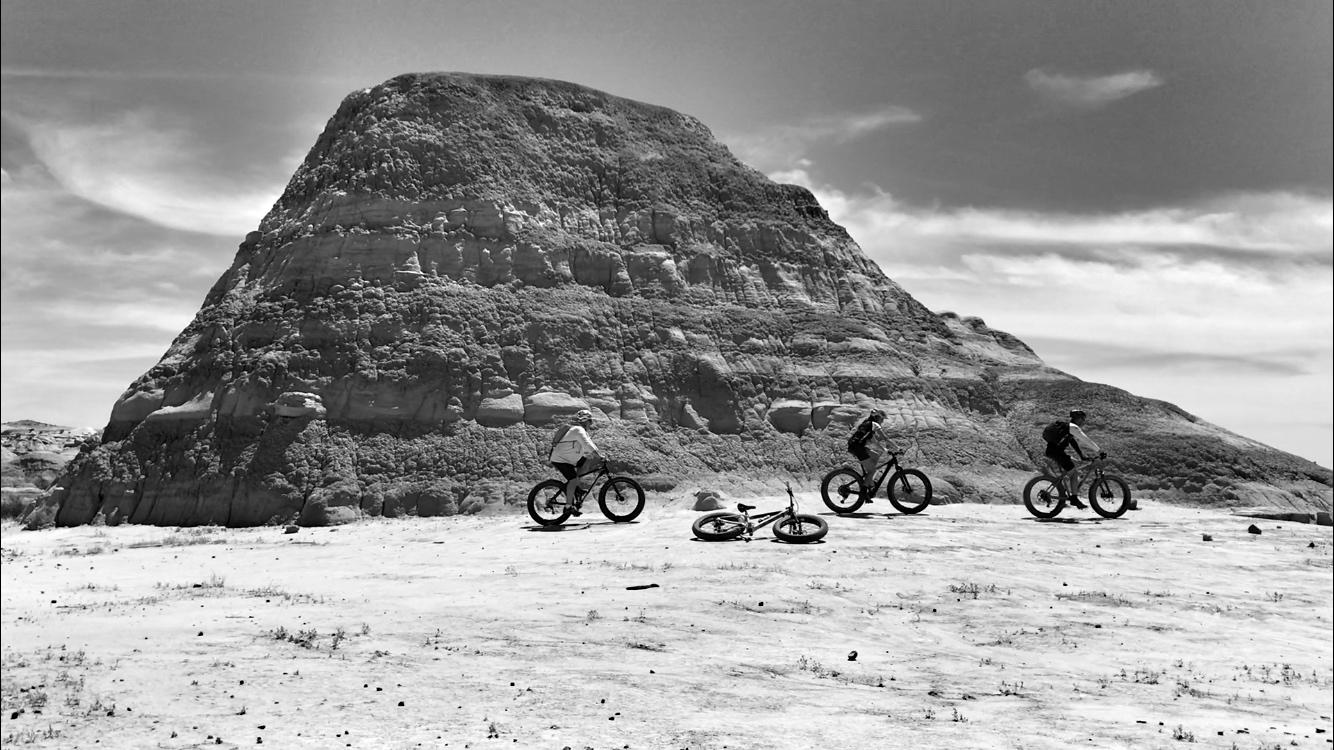 A group of four mountain bikers riding on a rocky terrain with a large, steep hill in the background. Two bikes are lying on the ground, and the scene is captured in black and white under a partly cloudy sky. Double Slot Fat Bike Trail mountain bike trail.