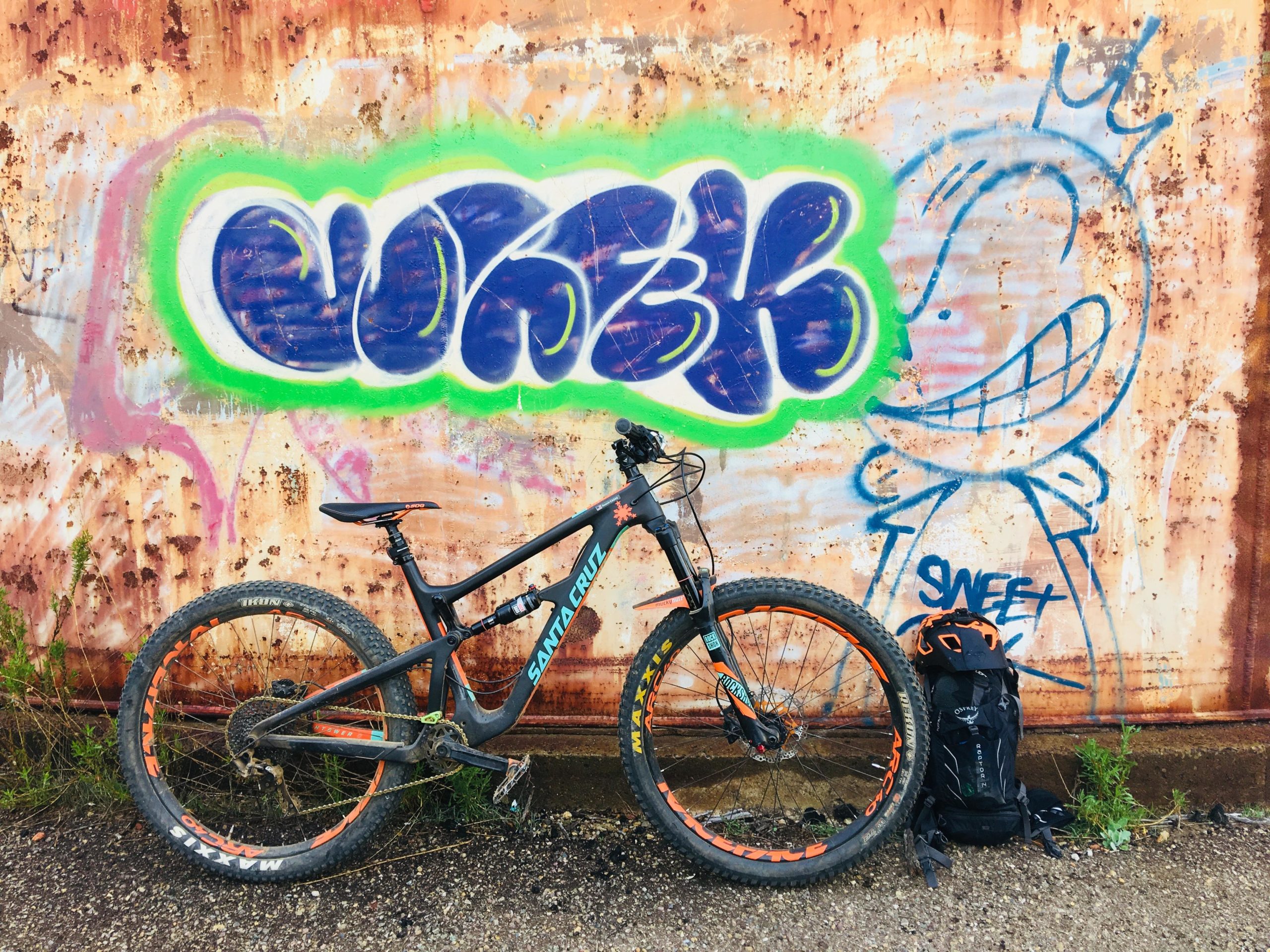 A mountain bike leaning against a rusted metal wall adorned with colorful graffiti. The bike, featuring a black frame and orange accents, is positioned next to a black backpack. The graffiti includes the words "SW3RK" and a cartoonish character with a big grin. The scene is set in an urban environment with some greenery visible at the base of the wall. Daley Ranch mountain bike trail.