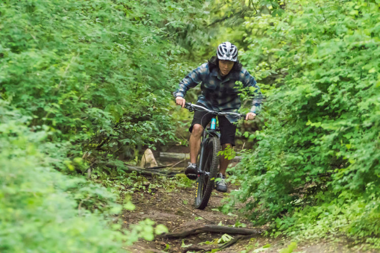 A mountain biker navigates a narrow, wooded trail surrounded by lush greenery, wearing a helmet and a plaid shirt. The rider is focused, demonstrating skill as they maneuver over the terrain.