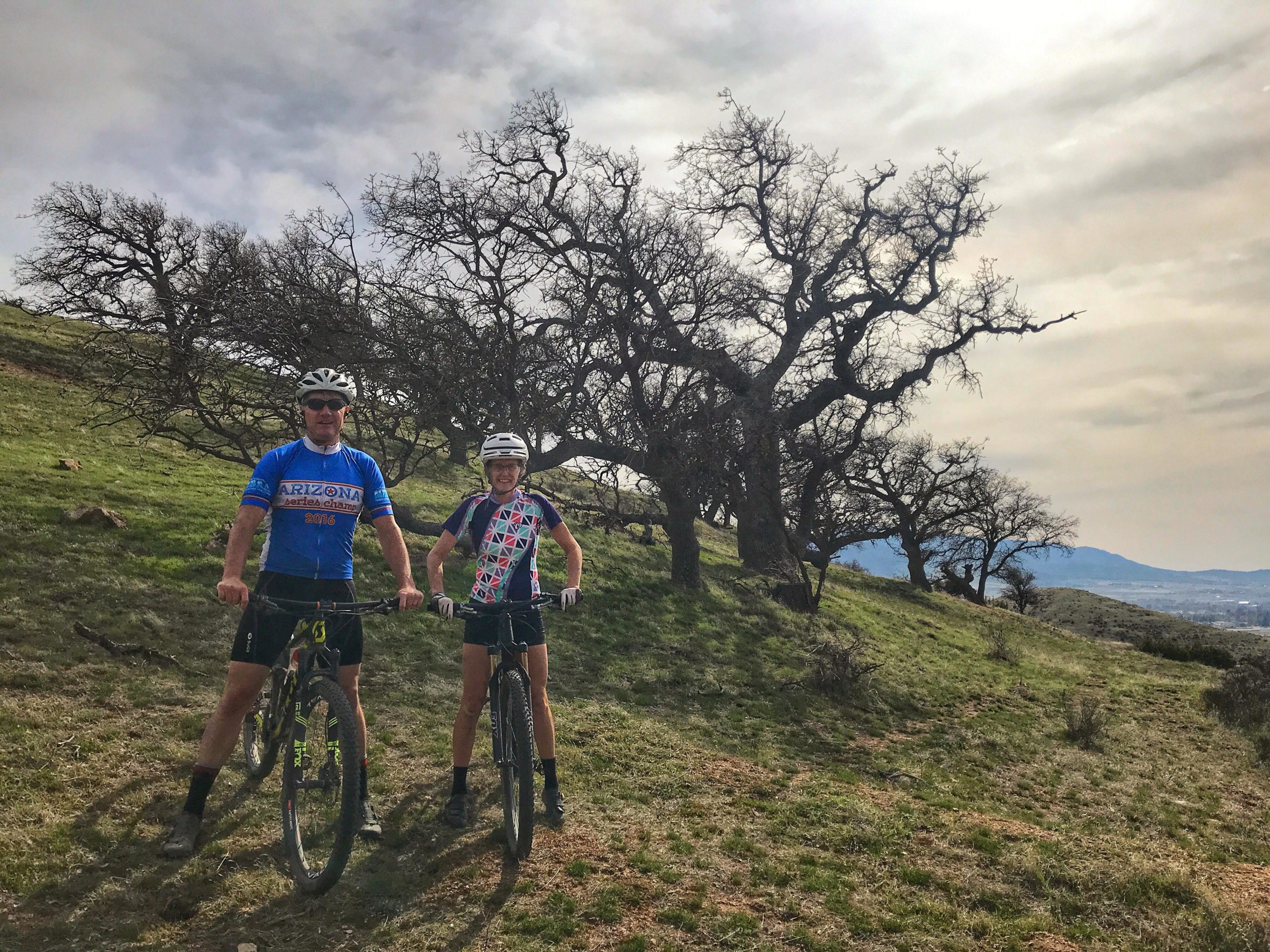 Two mountain bikers pose with their bikes on a grassy hill, surrounded by bare trees and a cloudy sky. They wear helmets and colorful jerseys, showcasing a serene outdoor setting perfect for biking. TMTA Lehigh trails mountain bike trail.