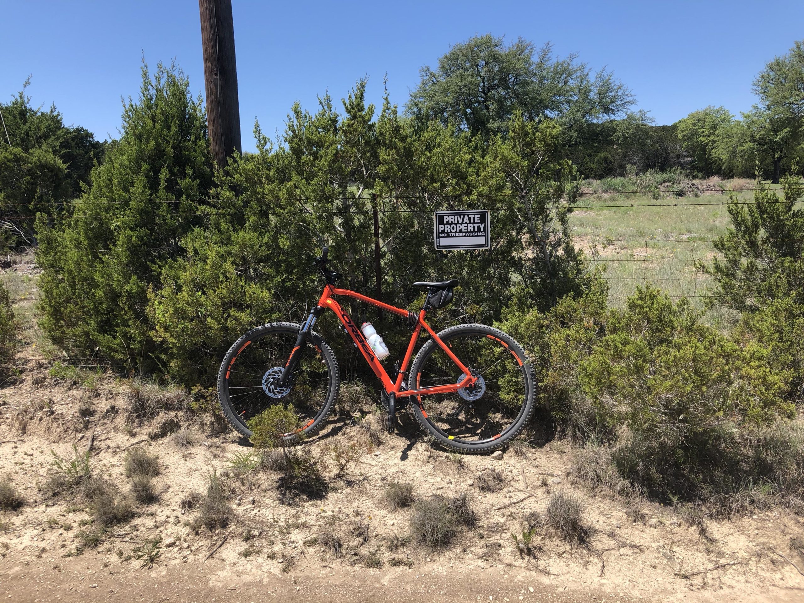 A bright orange mountain bike is leaning against a bush near a sign that reads "PRIVATE PROPERTY NO TRESPASSING." The scene is outdoors, showcasing a gravel road, green shrubs, and a clear blue sky. A wooden utility pole is visible in the background. Dana Peak mountain bike trail.