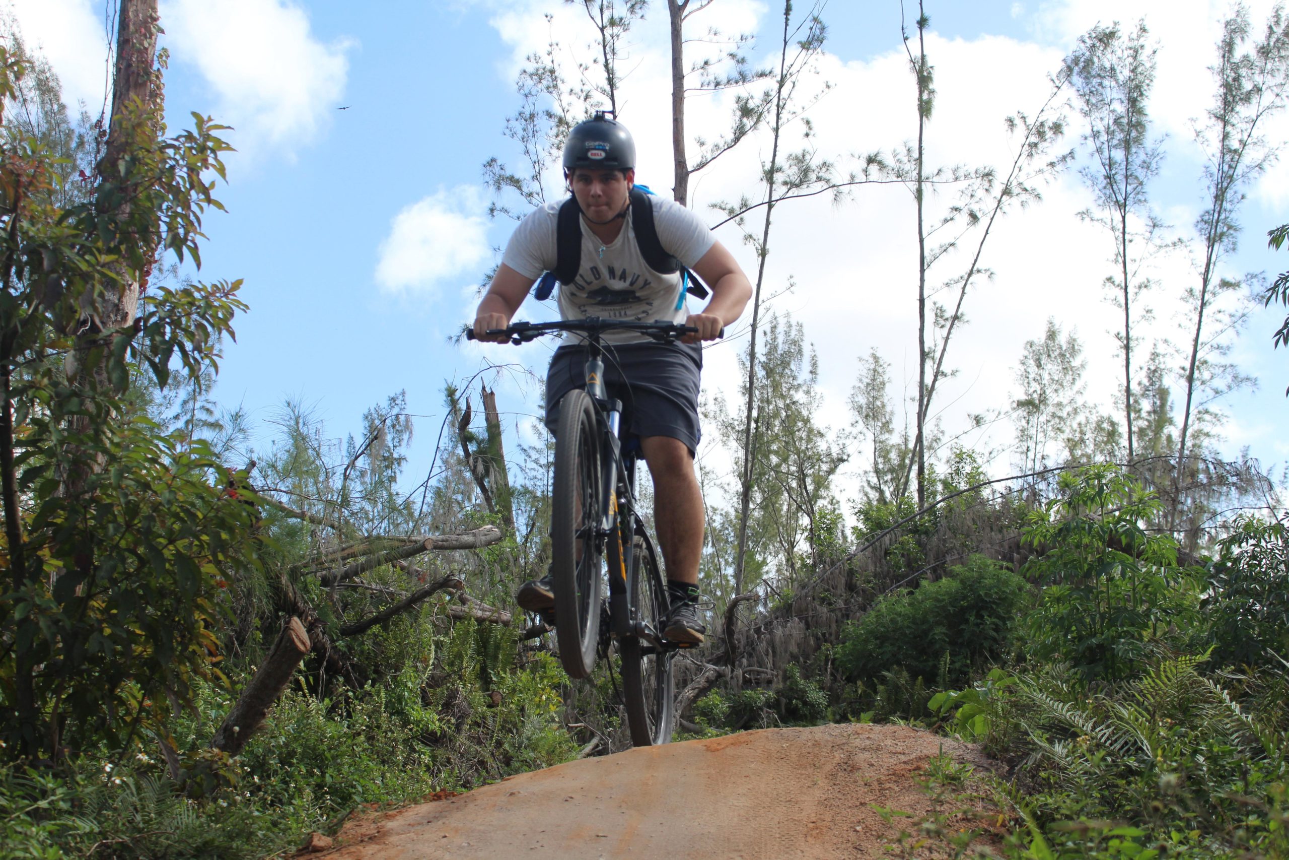 A cyclist performing a jump on a mountain bike on a dirt trail surrounded by greenery and fallen trees, under a partly cloudy sky. Amelia Earhart Park mountain bike trail.