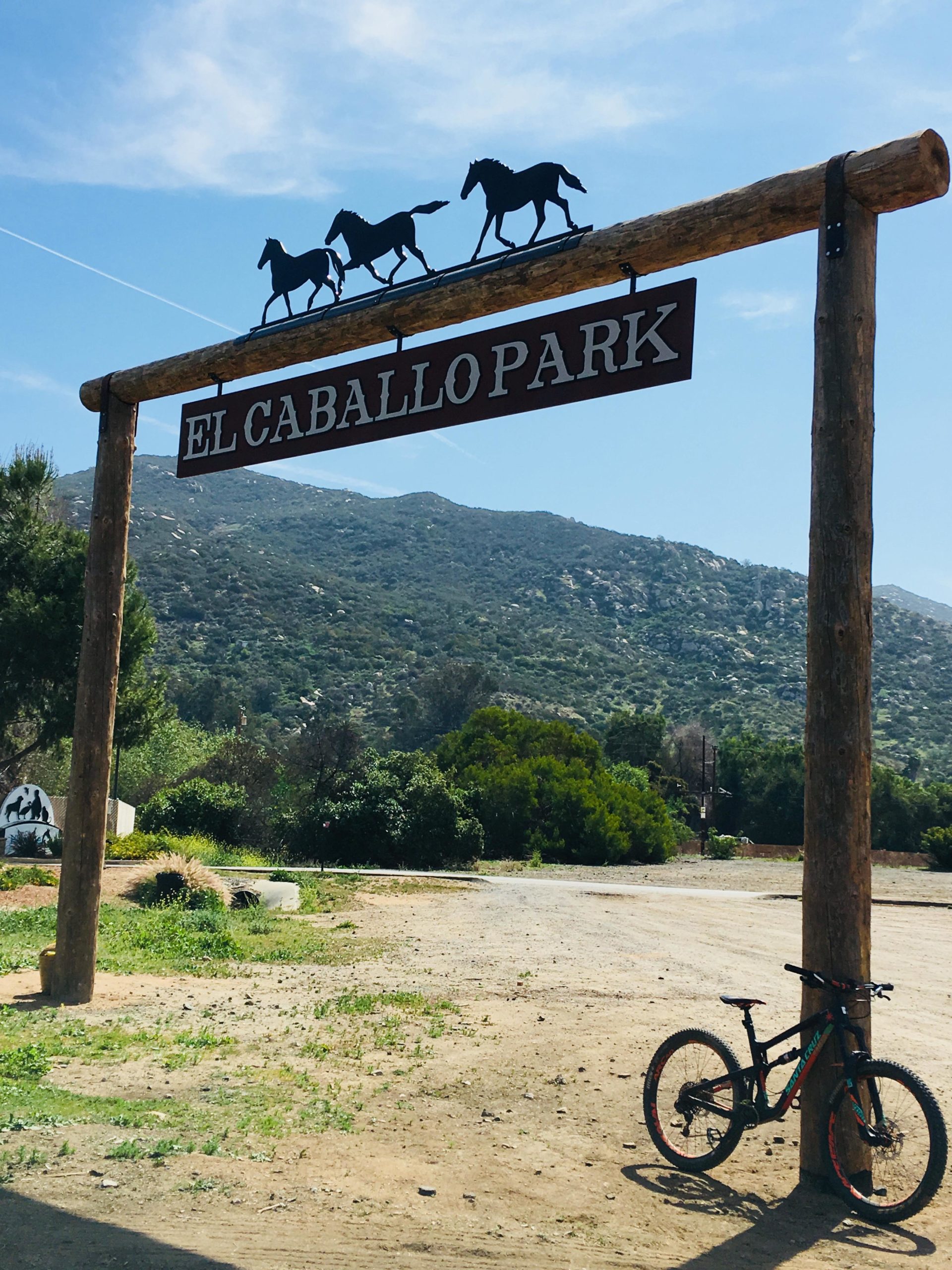 A wooden entrance sign with the text "El Caballo Park" and silhouettes of running horses on top, set against a backdrop of mountains and a clear blue sky. A bicycle leans against a post next to the sign, with a dirt path and greenery around. Daley Ranch mountain bike trail.