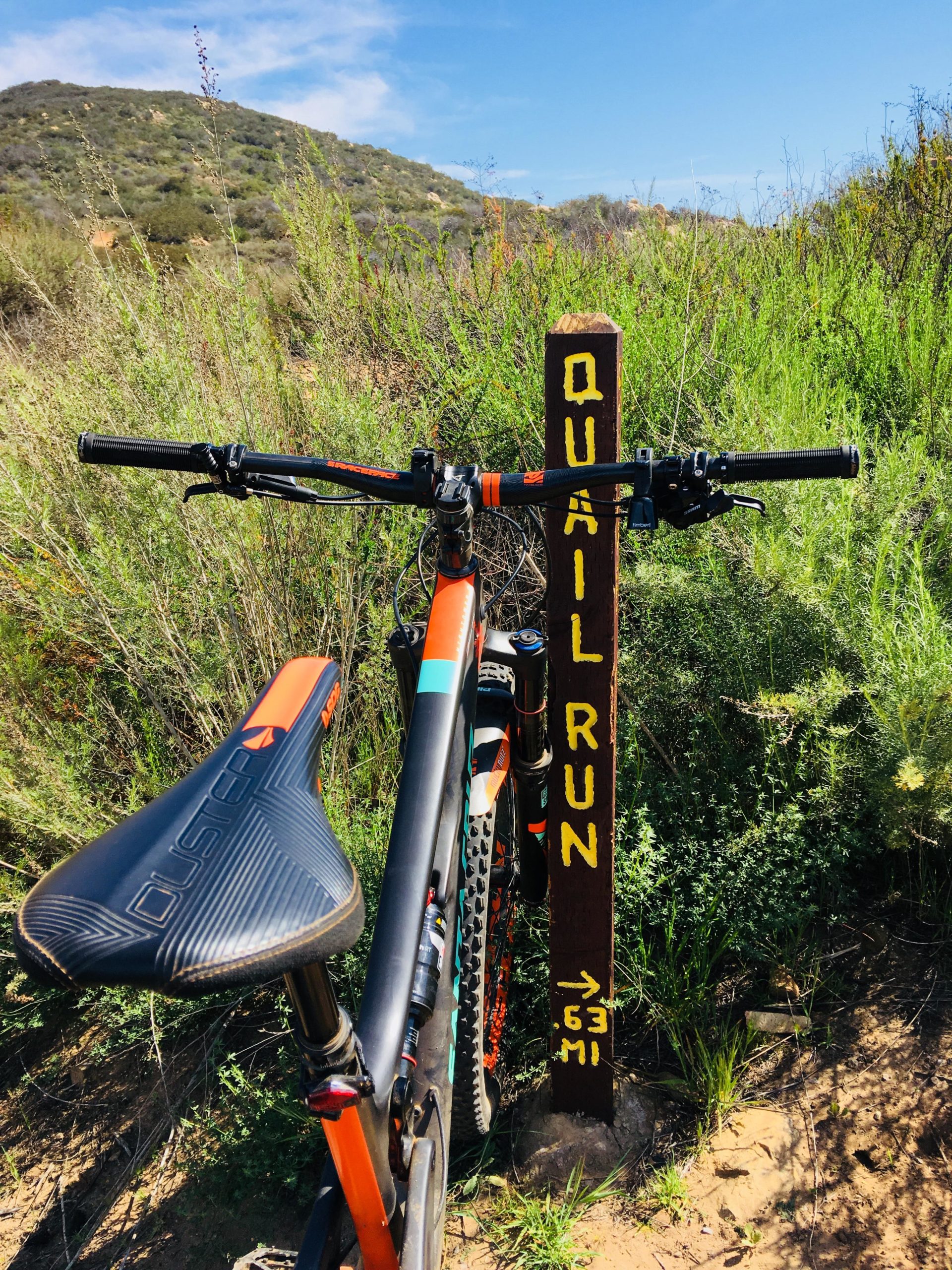 A mountain bike is positioned next to a wooden trail sign that reads "Quail Run → 0.63 mi," surrounded by green vegetation and hills in the background under a clear blue sky. Daley Ranch mountain bike trail.