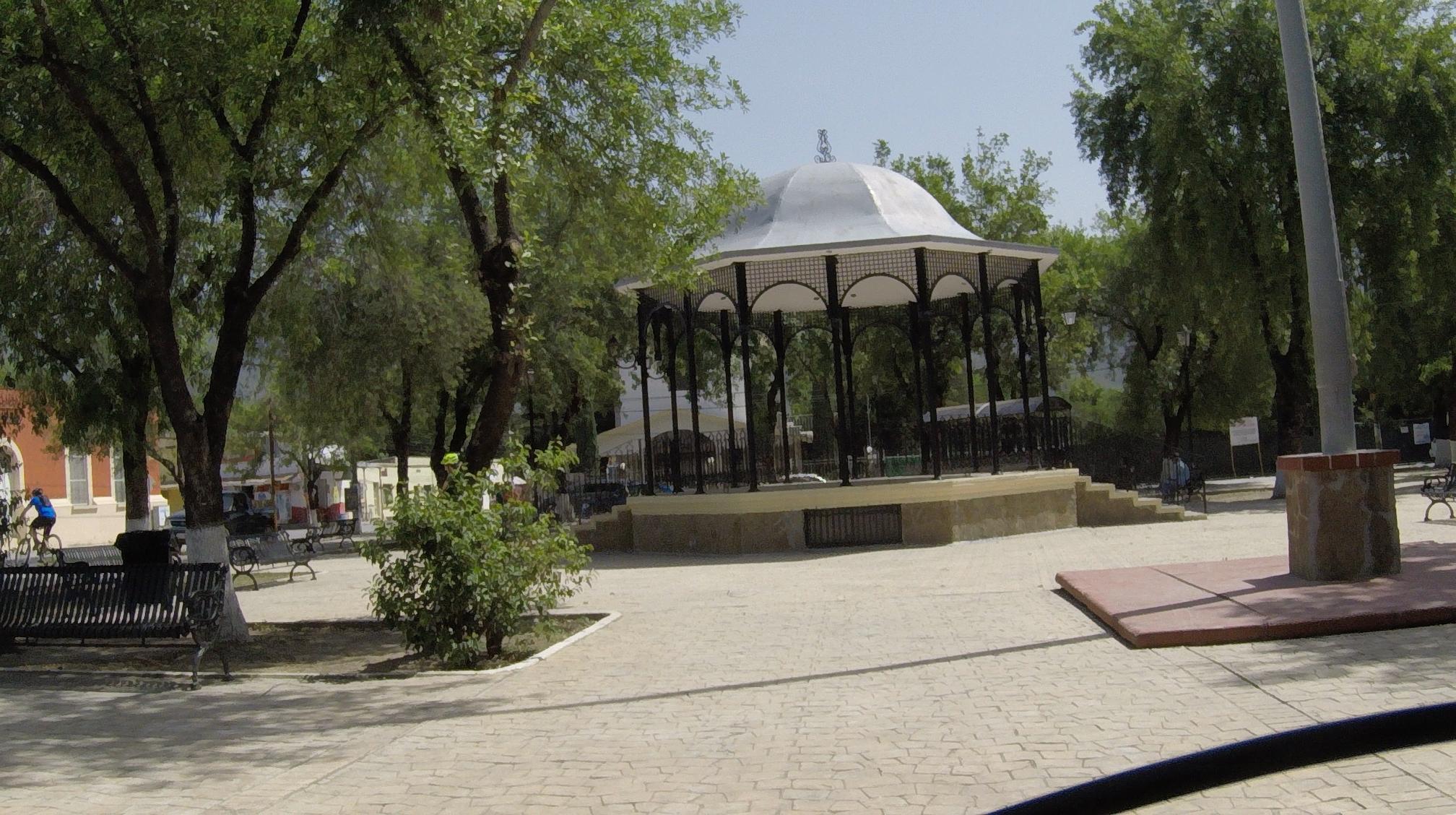 A tranquil park scene featuring a gazebo surrounded by trees and benches. In the background, a person on a bicycle is visible, and the paved walkway leads up to the gazebo's steps, creating a peaceful atmosphere. Bosque Encantado mountain bike trail.