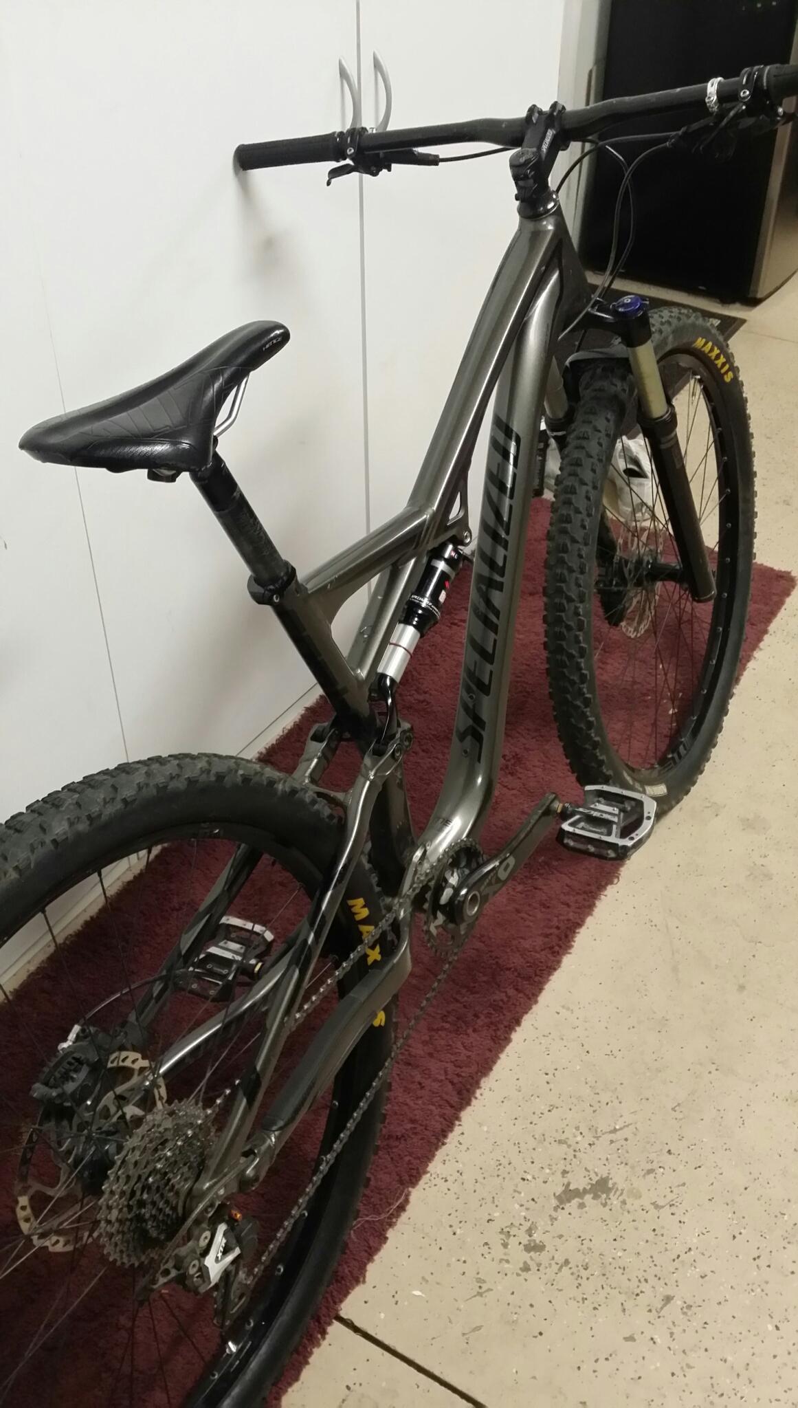 Specialized Camber 29: A gray mountain bike leaning against a white wall, featuring thick tires and a black saddle. The bike has visible gears and suspension, with the brand name "Specialized" displayed prominently on the frame. It rests on a patterned rug in a garage or indoor space, with additional household items blurred in the background.
