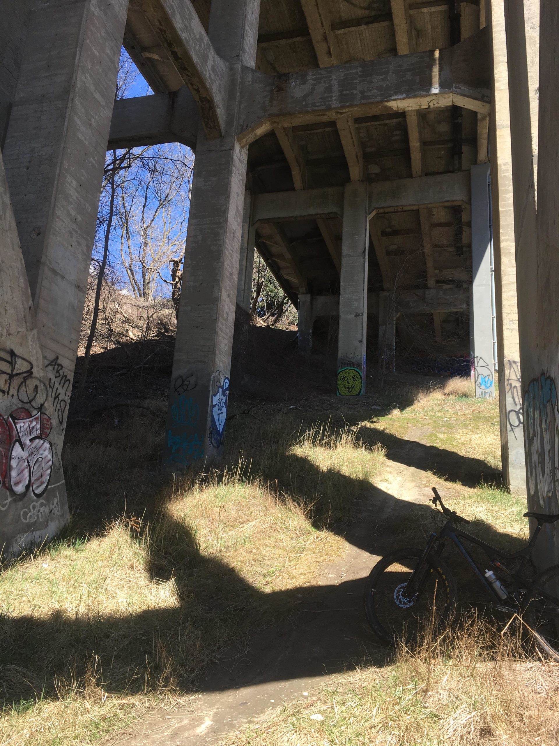 A view of a shaded area beneath a concrete overpass, featuring large pillars covered in graffiti. Sunlight filters in from above, illuminating a dirt path lined with grassy patches. A black mountain bike rests on the ground beside the path. Don Valley mountain bike trail.
