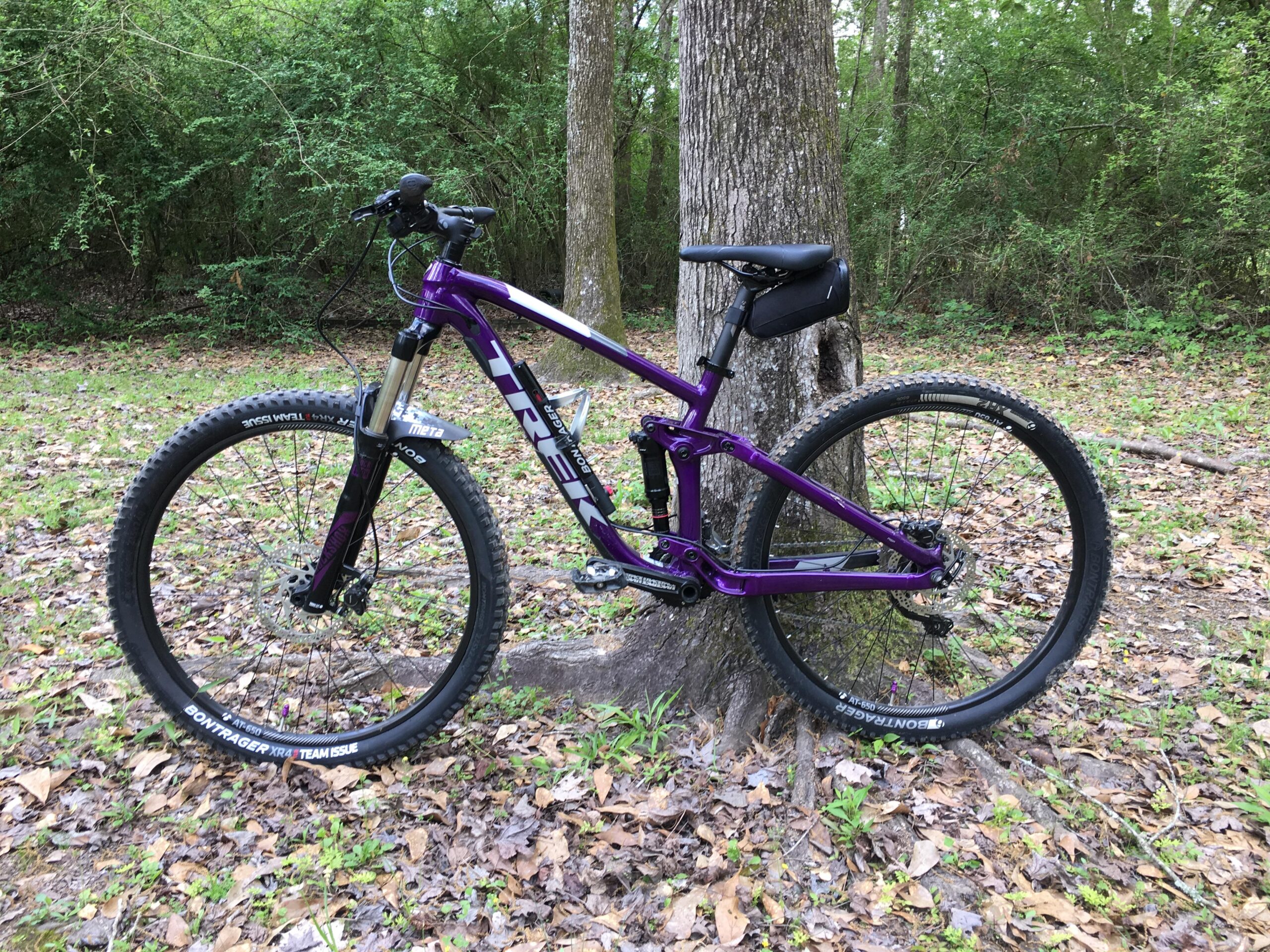 Trek Fuel EX 5: A purple mountain bike leaning against a tree, set in a natural environment with scattered leaves and greenery in the background. The bike features thick tires and a sturdy frame, designed for off-road riding.