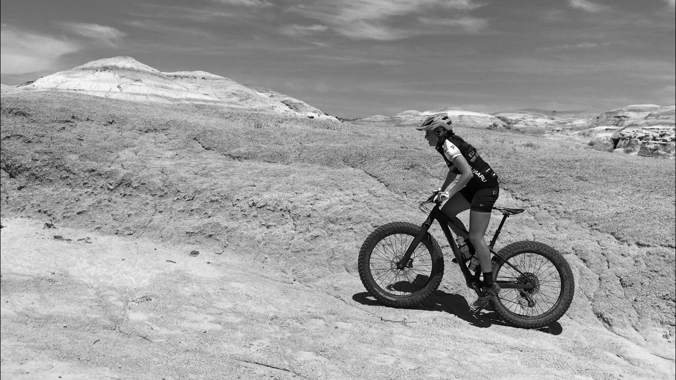 A person riding a fat tire mountain bike on a rocky, uneven terrain under a bright sky, captured in black and white. The cyclist appears focused, dressed in athletic gear, and is navigating the challenging landscape. Double Slot Fat Bike Trail mountain bike trail.