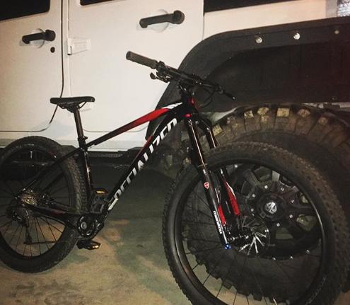 Specialized FUSE EXPERT 6FATTIE: A black and red mountain bike is parked in a garage beside a rugged vehicle with large tires. The bike is leaning against the tire of the vehicle, and the background shows part of the car's body. The setting appears dimly lit, emphasizing the bike's sleek design.