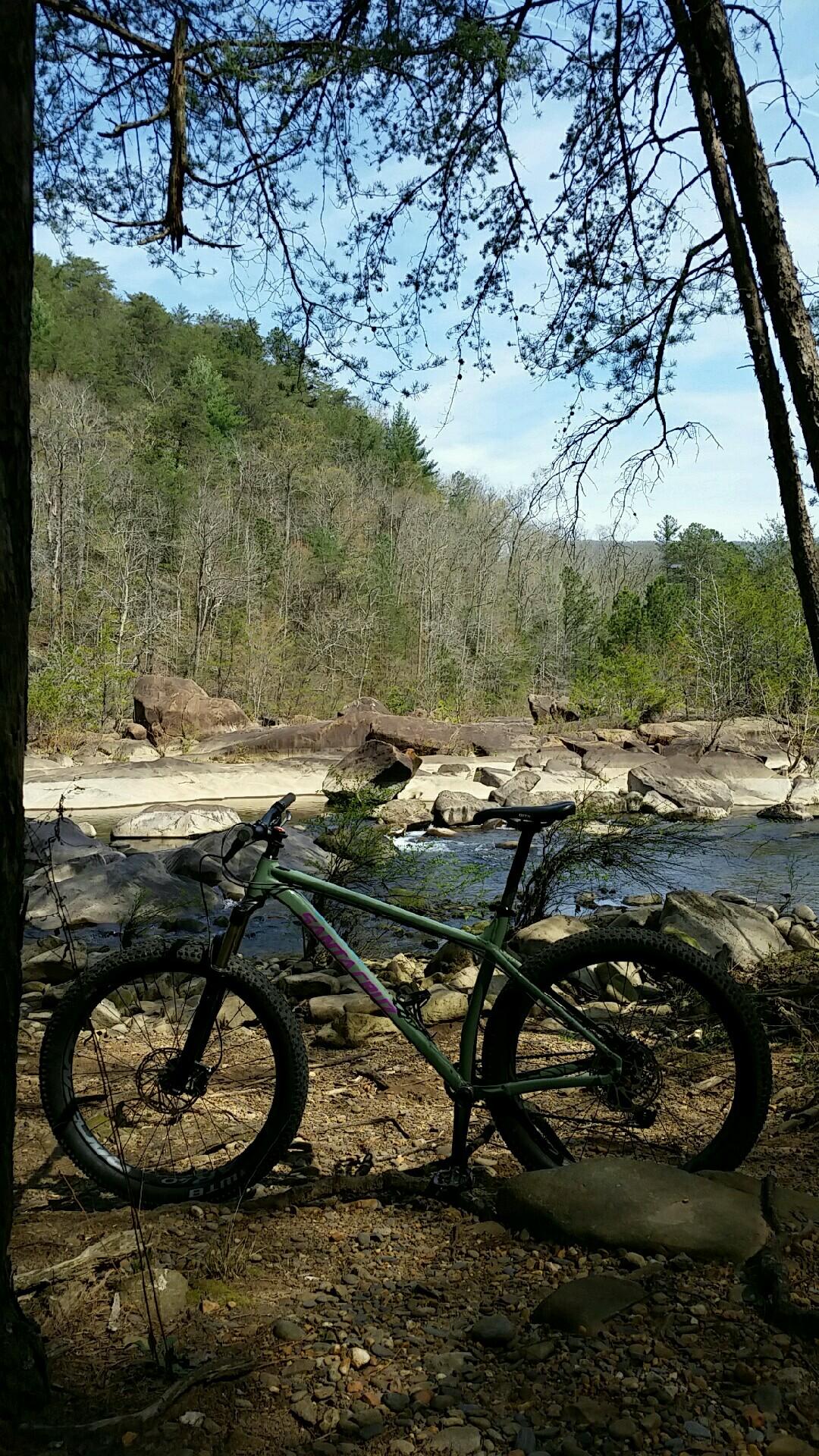 A mountain bike resting on a rocky riverbank surrounded by trees and boulders. The scene features a clear blue sky with scattered clouds, and the flowing river can be seen in the background. Old Copper Rd mountain bike trail.