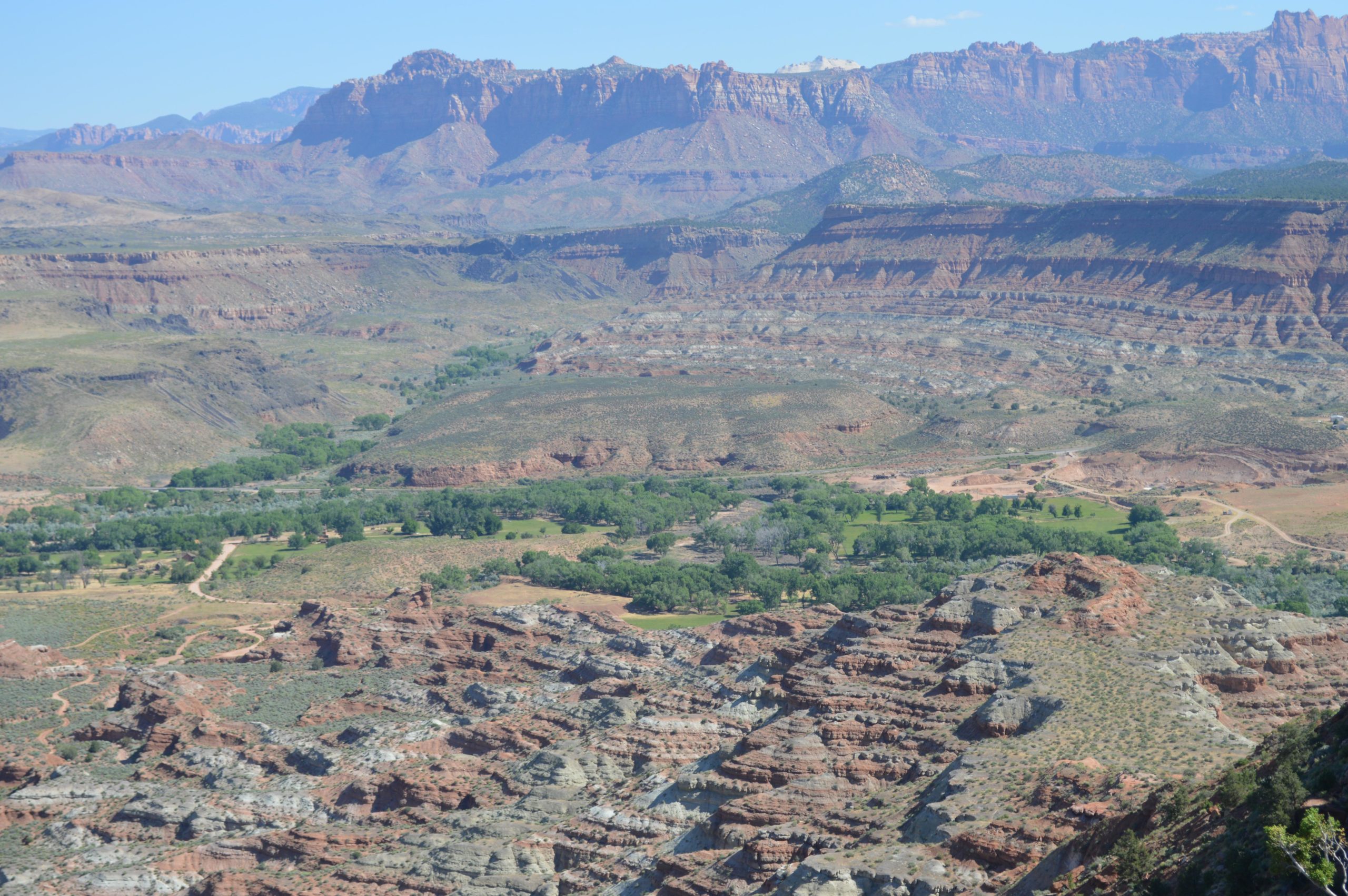 A panoramic view of a rugged landscape featuring layered rock formations and valleys, with patches of green vegetation interspersed throughout. The backdrop consists of distant mountains under a clear blue sky, highlighting the natural beauty of the terrain. Wire Mesa Loop mountain bike trail.