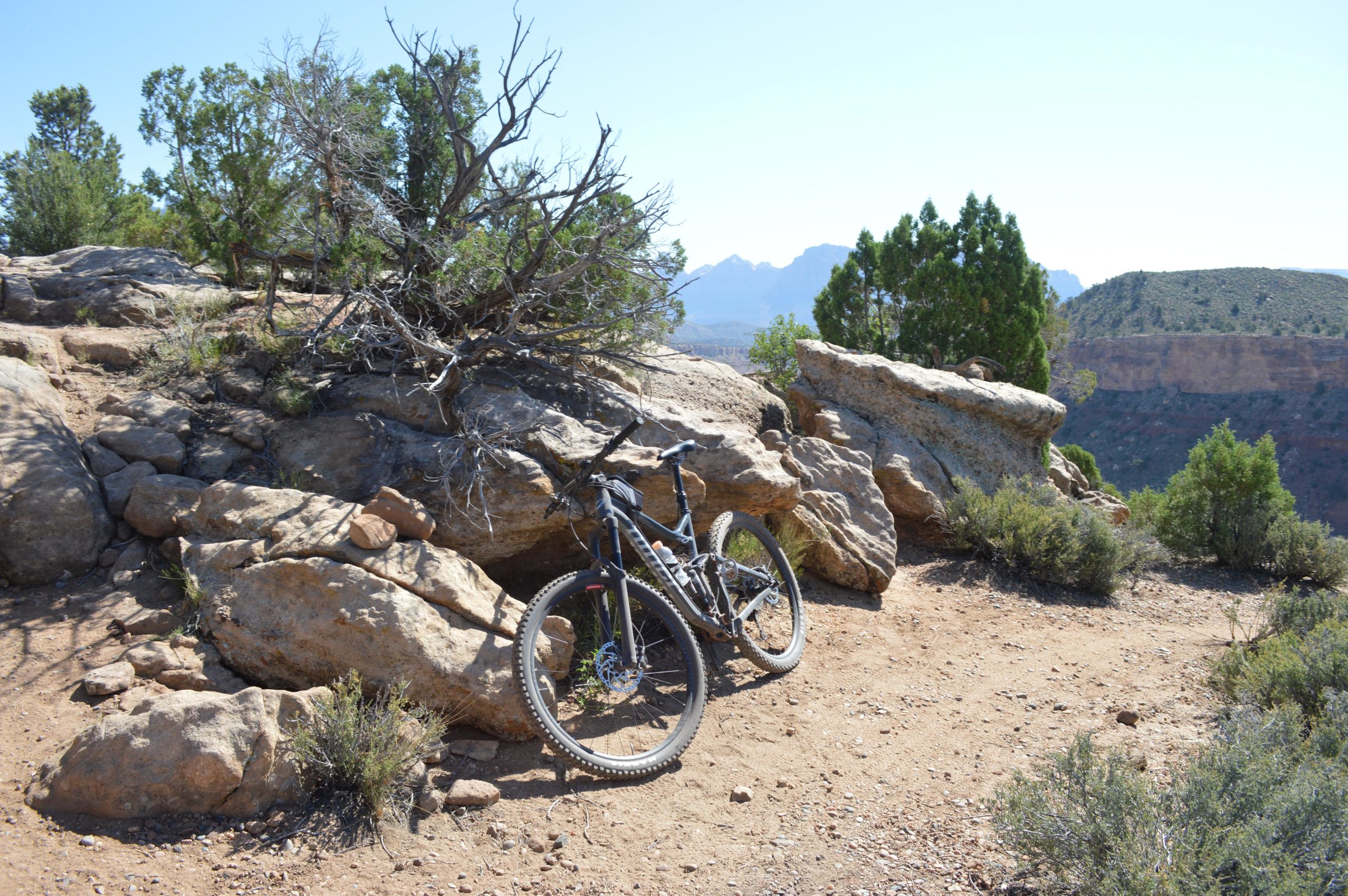 A mountain bike resting against a large rock outcropping in a rugged outdoor landscape, surrounded by shrubs and trees, with distant mountains visible in the background under a clear blue sky. Wire Mesa Loop mountain bike trail.
