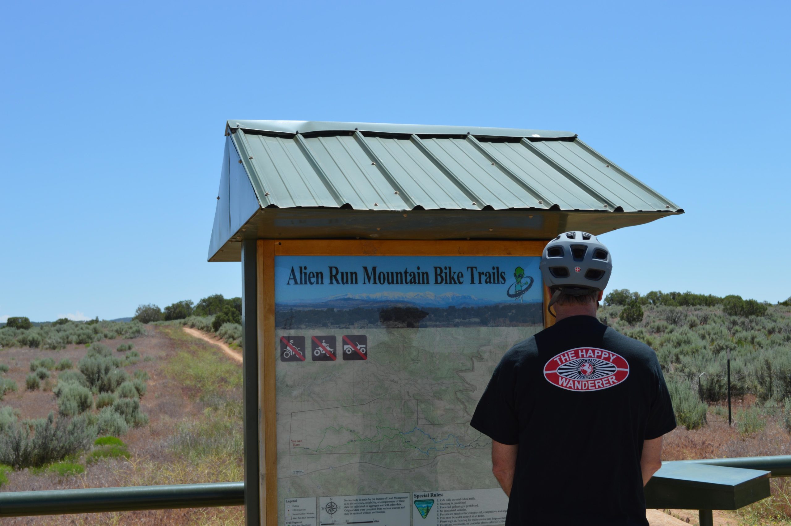 A person wearing a helmet stands in front of a mountain bike trail map at Alien Run Mountain Bike Trails. The map displays a trail overview and icons indicating trail rules. The background features a dirt path and open landscape with shrubs, under a clear blue sky. Alien Run Trail mountain bike trail.