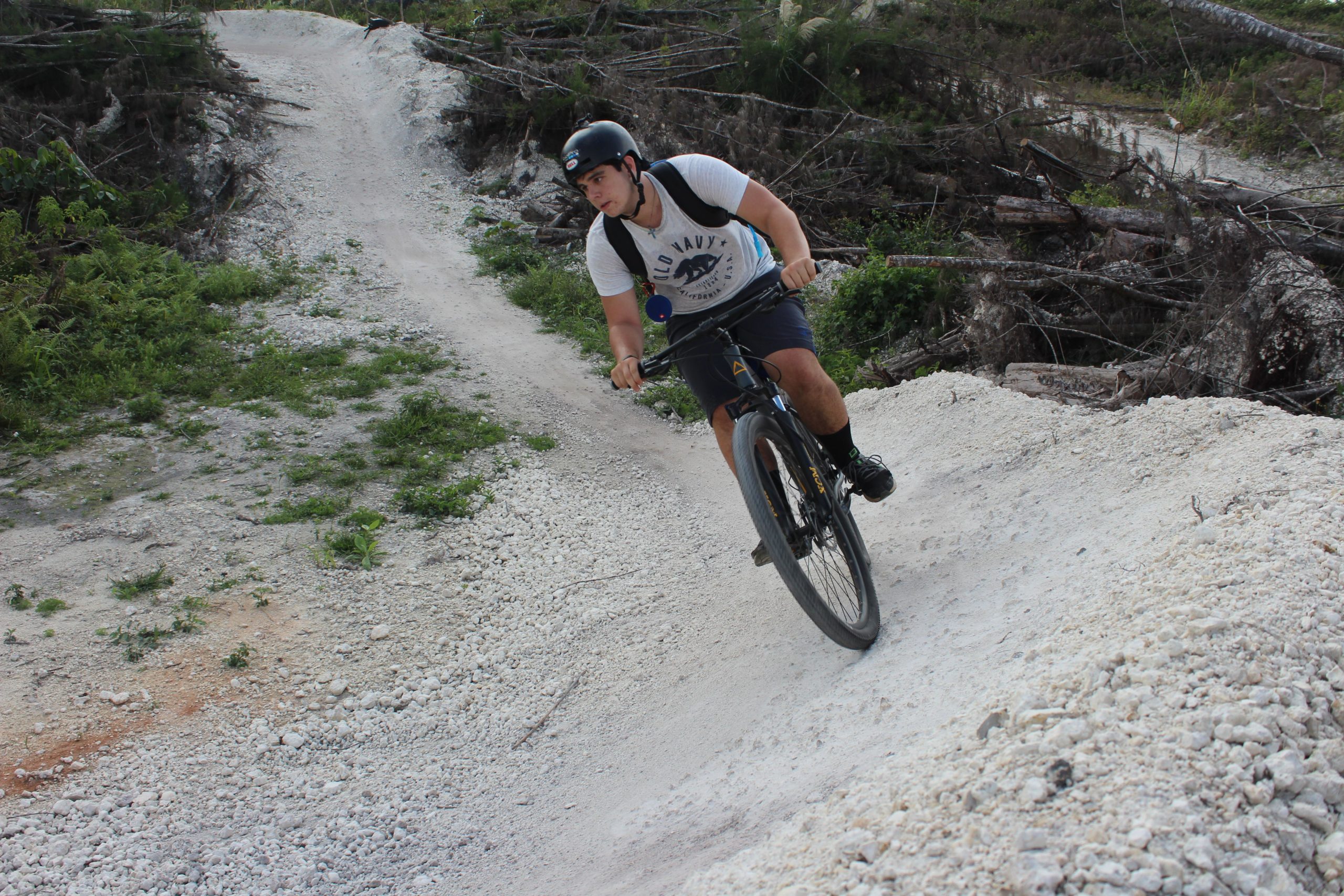 A person in casual clothing rides a mountain bike on a dirt trail surrounded by greenery and fallen tree debris. The individual is wearing a helmet and appears to be navigating a curve on the trail, showcasing an active outdoor setting. Amelia Earhart Park mountain bike trail.