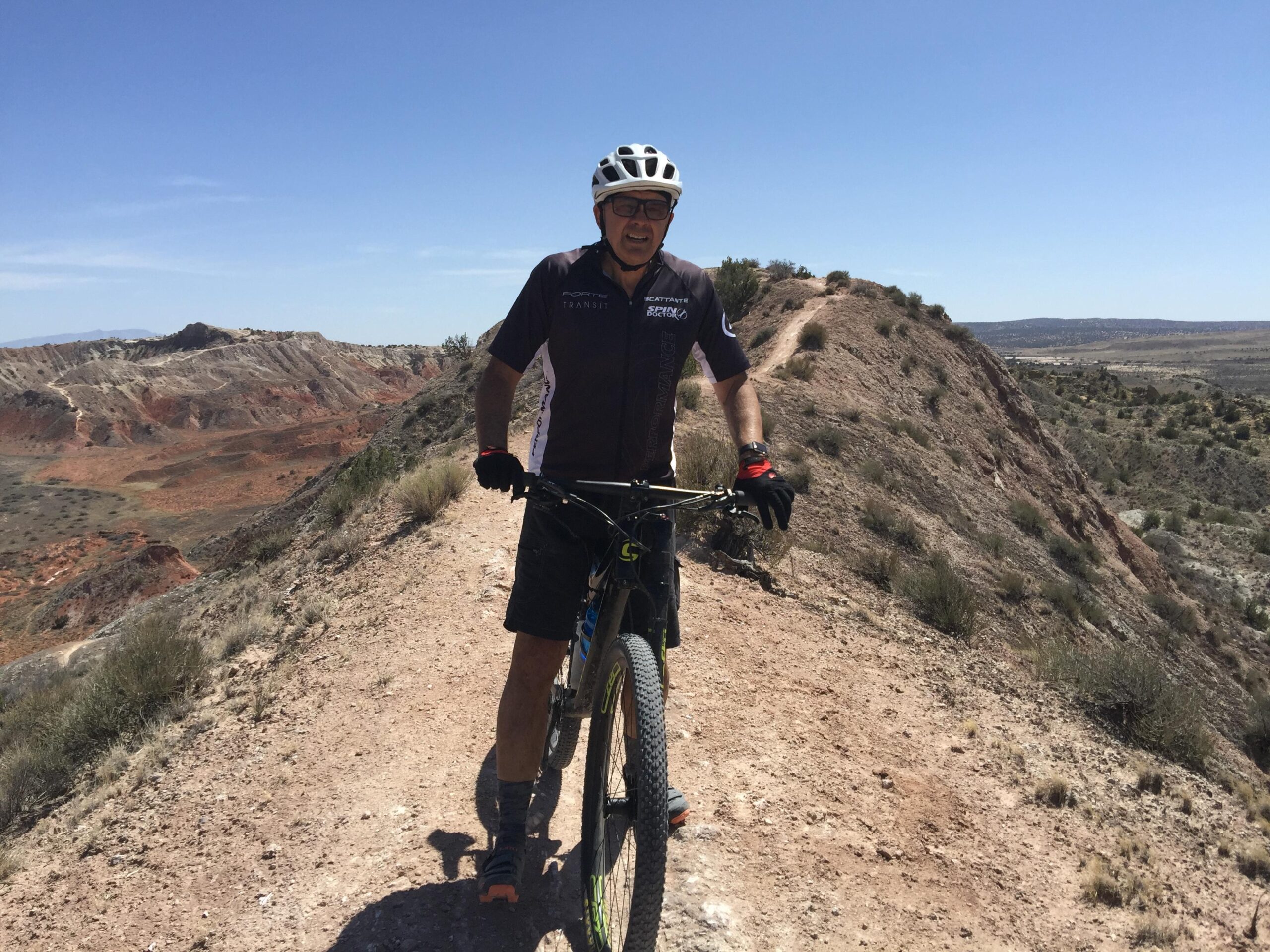 Cannondale Scalpel 29 Carbon 2: A mountain biker standing on a dirt trail with a rocky landscape in the background. The rider is wearing a helmet and cycling gear, with a sunny blue sky overhead. The terrain features hills and valleys characterized by reddish soil and sparse vegetation.