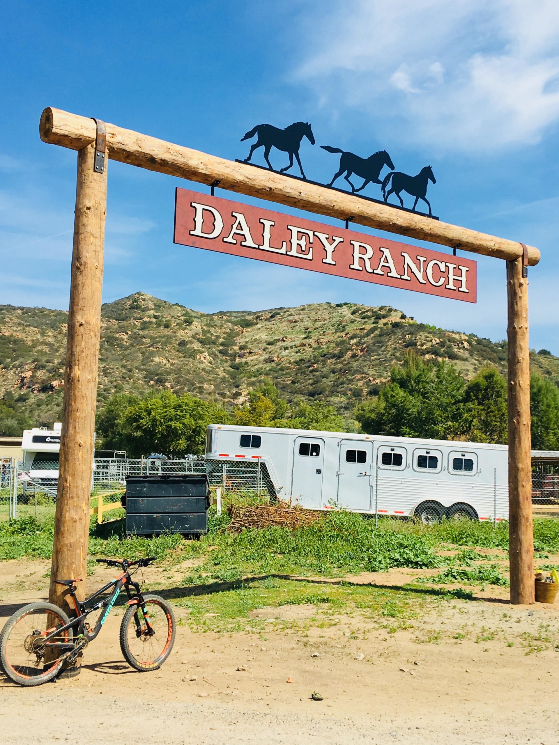 A rustic wooden sign featuring the words "Daley Ranch," with silhouettes of horses above it. In the foreground, a mountain bike leans against a post. Behind the sign, green hills can be seen, along with a horse trailer and various shrubs. The sky is blue with scattered clouds. Daley Ranch mountain bike trail.
