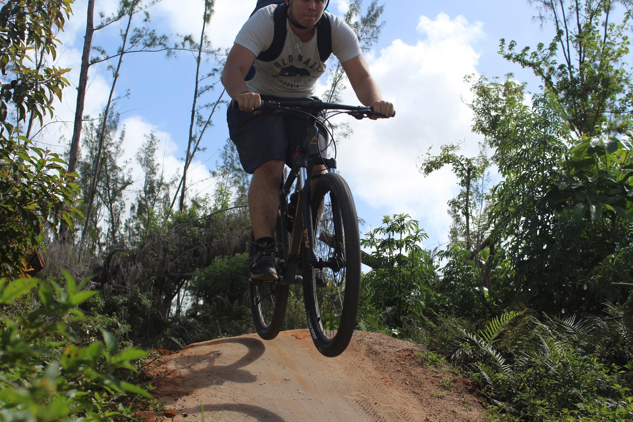 A person riding a mountain bike mid-jump over a dirt ramp, surrounded by lush greenery and trees under a partly cloudy sky. Amelia Earhart Park mountain bike trail.