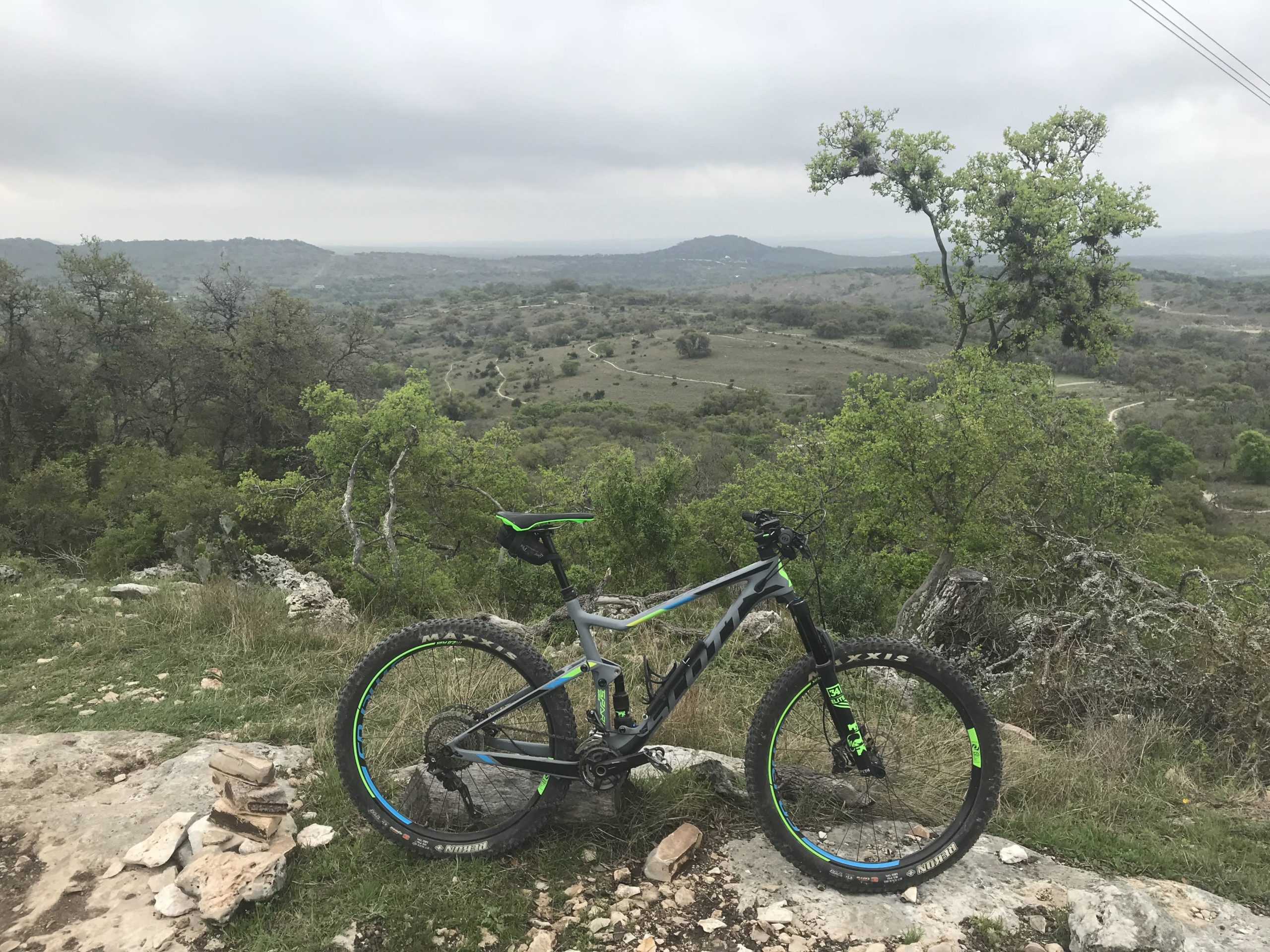 A mountain bike parked on rocky terrain, with a scenic view of rolling hills and greenery in the background. The sky is overcast, creating a moody atmosphere. Flat Rock Ranch mountain bike trail.