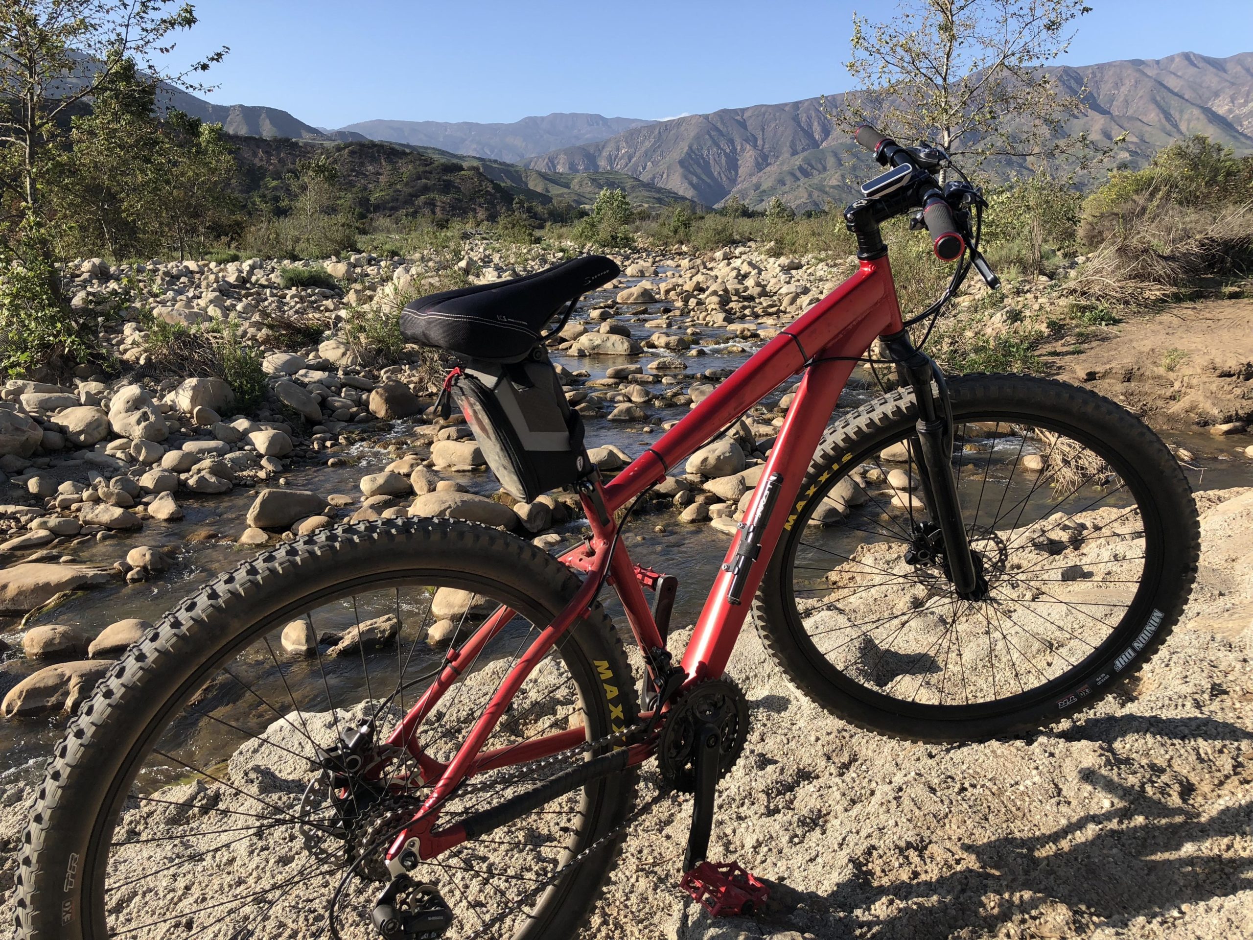 A red mountain bike parked on a rocky shore beside a stream, with rolling hills and mountains in the background under a clear blue sky. The bike