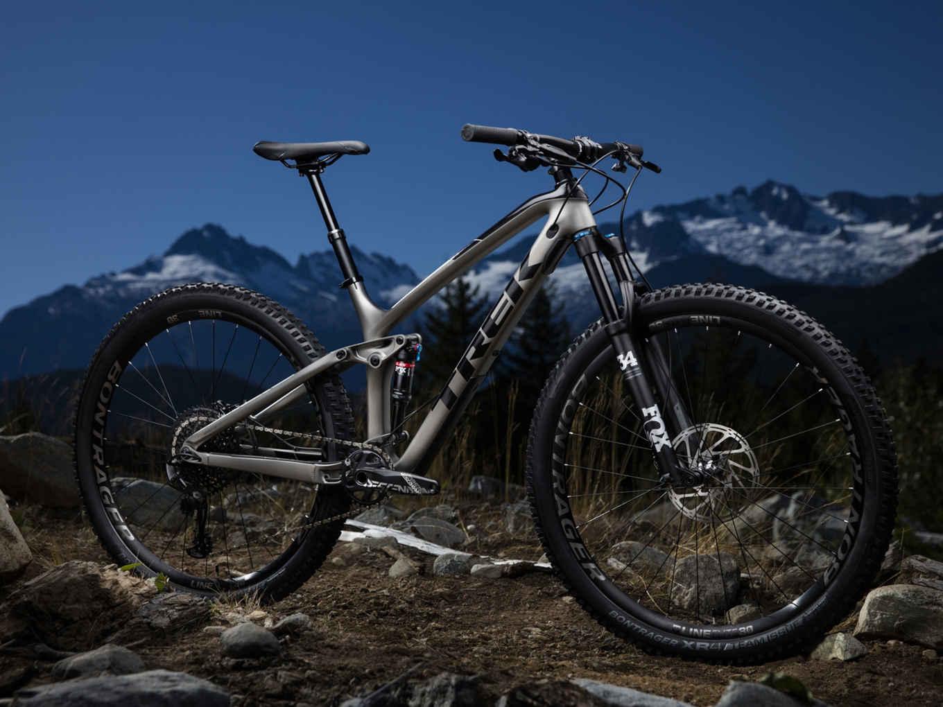 Trek Fuel EX 9.8: A mountain bike positioned on rocky terrain with a scenic backdrop of snow-capped mountains under a blue sky. The bike features a silver frame, prominent front suspension, and rugged tires designed for off-road use.