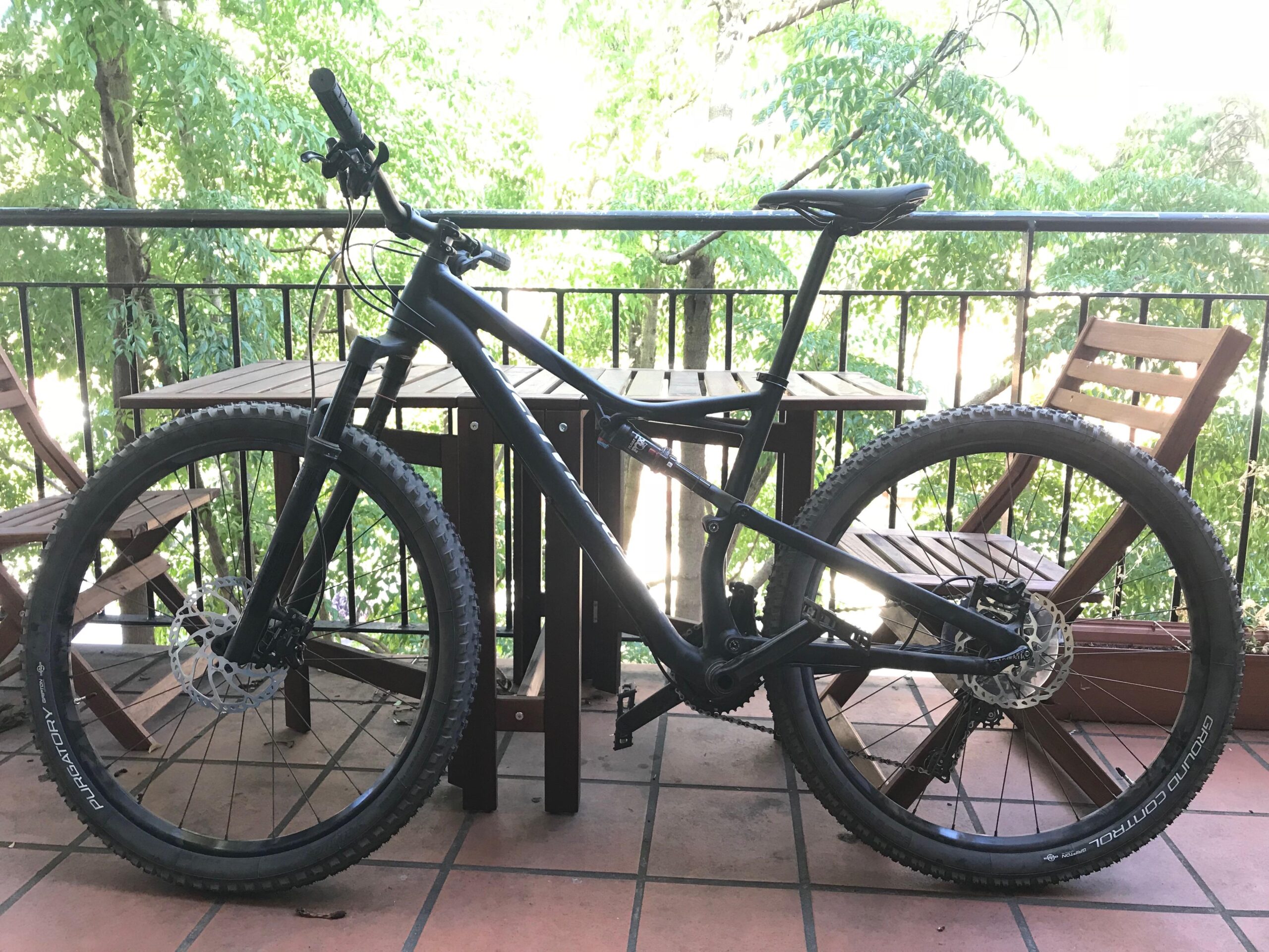 Specialized Camber Comp 29: A black mountain bike is positioned on a balcony with a wooden table and chair visible in the background. The bike features thick tires and a sturdy frame, with lush green trees visible behind it, suggesting a natural outdoor setting.