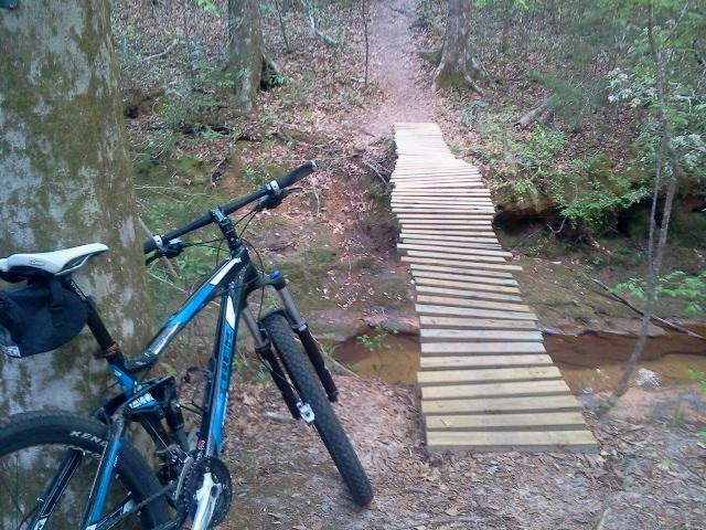 Trek Fuel EX 5: A mountain bike resting against a tree near a wooden bridge that crosses a small creek in a wooded area. A dirt path continues beyond the bridge, surrounded by lush greenery and fallen leaves.