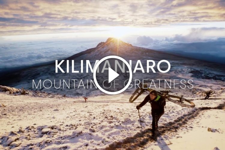 A hiker carrying a backpack and trekking poles ascends the snowy slopes of Kilimanjaro, with dramatic clouds and sunlight illuminating the mountain in the background. The text "Kilimanjaro - Mountain of Greatness" is overlaid on the image, featuring a play button icon, indicating that the image links to a video.
