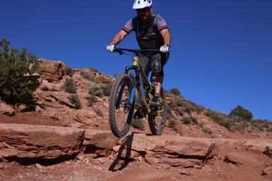 A mountain biker in a helmet performs a jump over a rocky terrain against a clear blue sky, showcasing an action-packed moment on a trail. Porcupine Rim mountain bike trail.