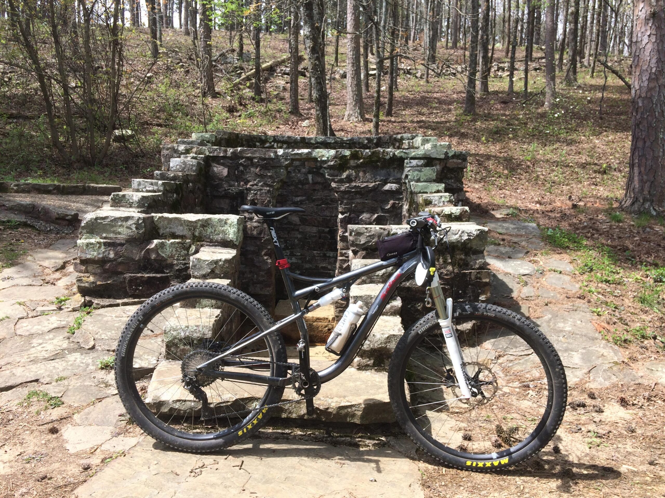 Salsa Horsethief: A mountain bike parked beside a stone structure in a wooded area. The bike has thick tires and suspension, and the surrounding landscape features trees and forest floor foliage.