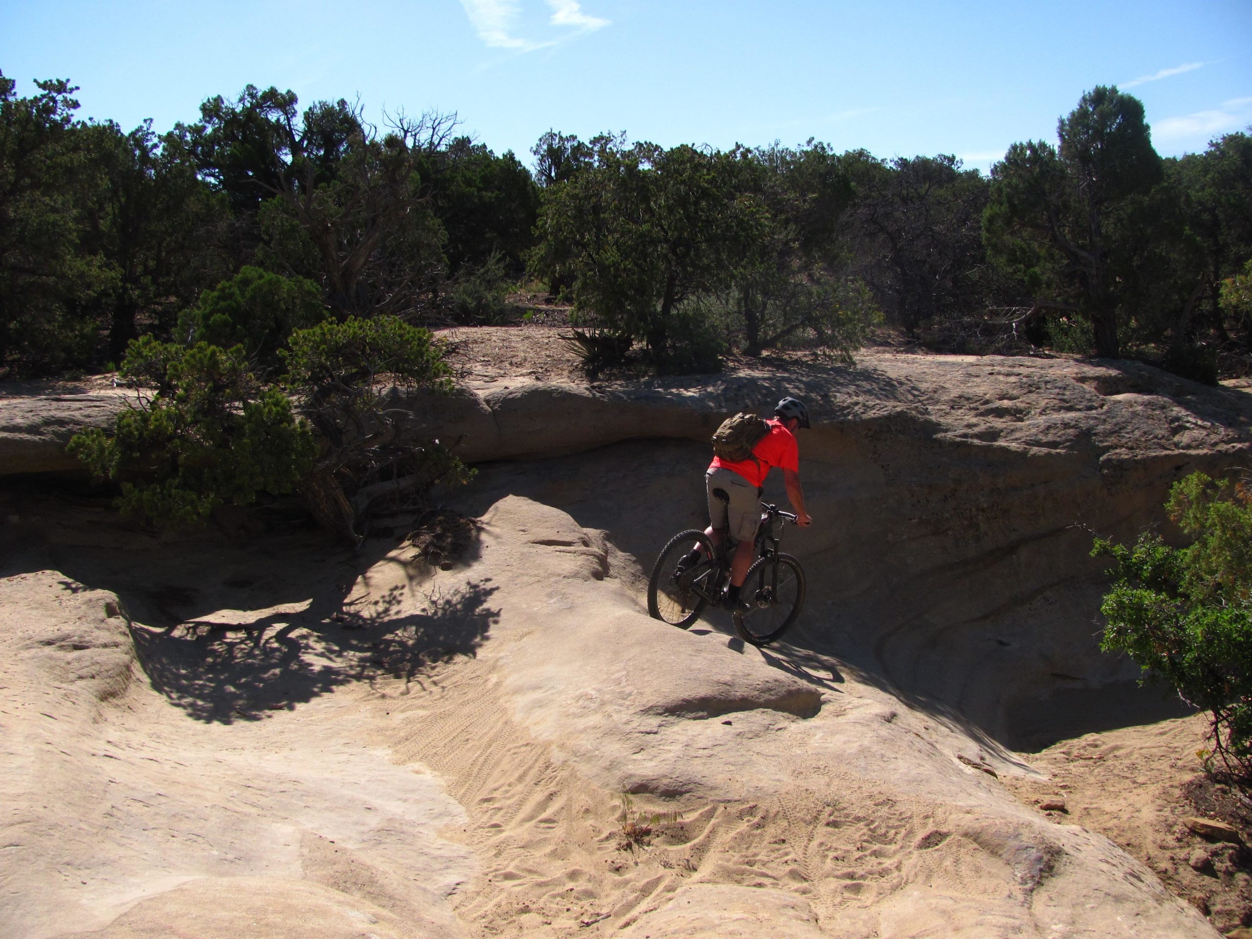 A mountain biker wearing a bright orange shirt and a helmet rides along a rocky trail surrounded by sparse vegetation and trees under a clear blue sky. The terrain is uneven, with a visible incline and sandy patches. Alien Run Trail mountain bike trail.