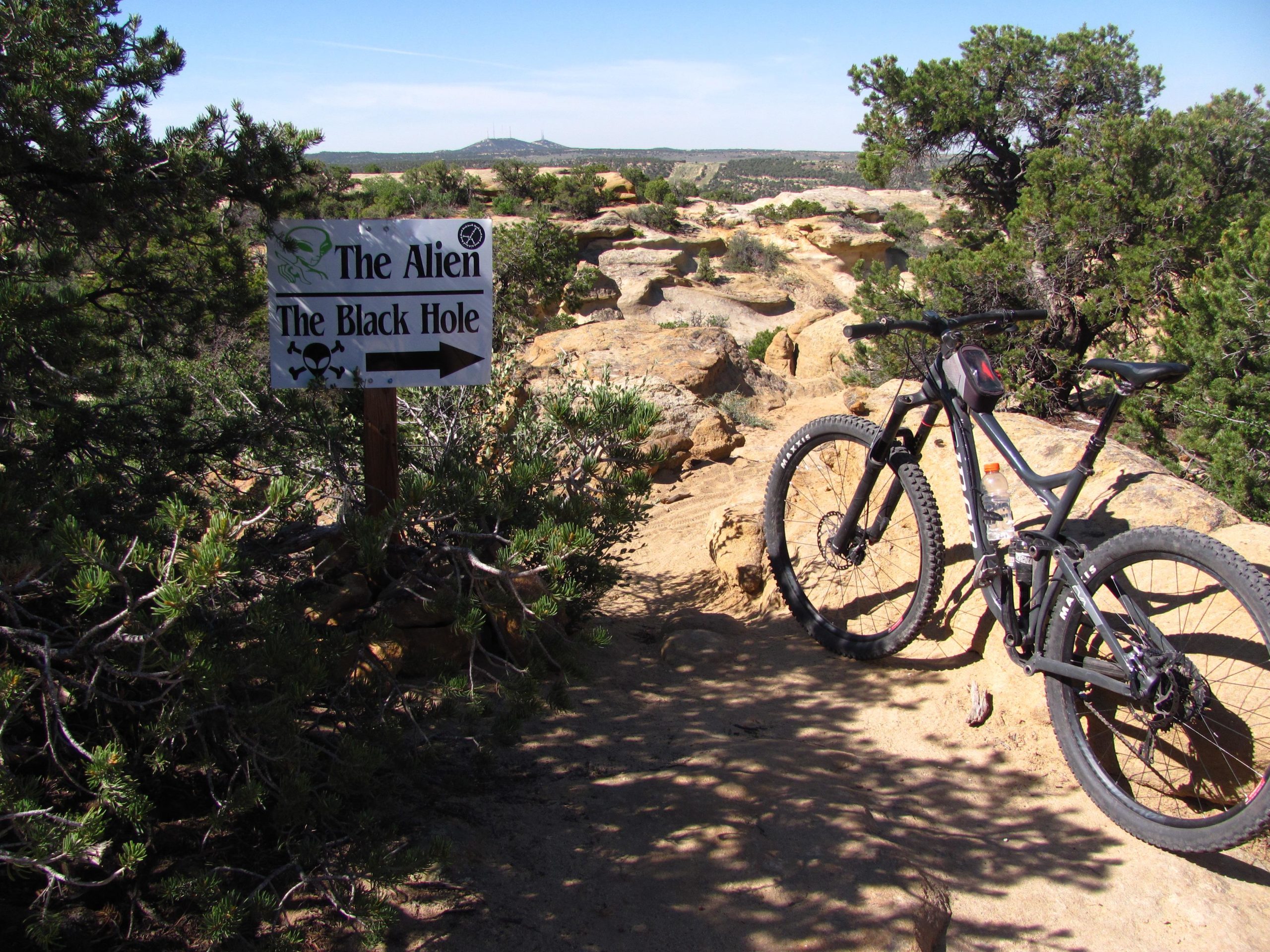 A mountain bike parked near a trail sign directing to "The Alien" and "The Black Hole," set in a rocky landscape with sparse greenery and a clear blue sky in the background. Alien Run Trail mountain bike trail.