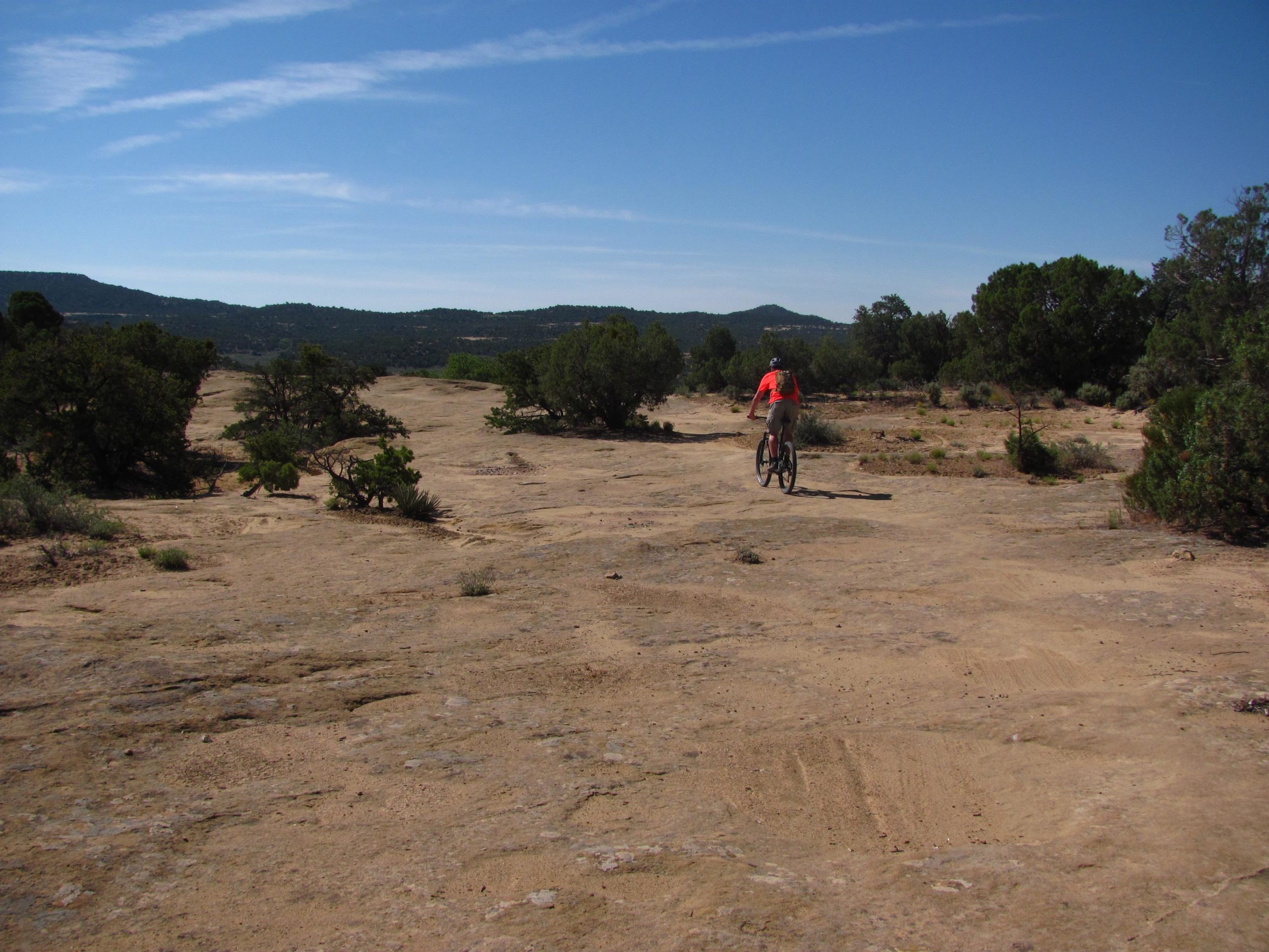 A person riding a mountain bike on a rocky trail, surrounded by sparse vegetation and scenic hills under a clear blue sky. Alien Run Trail mountain bike trail.