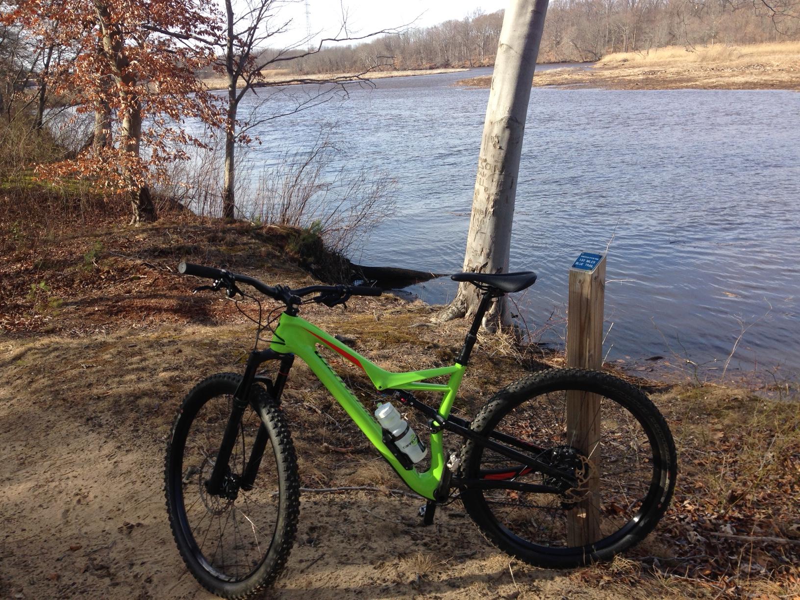 A bright green mountain bike leaning against a wooden post near a riverbank, surrounded by trees and dry leaves. The calm river reflects the clear sky, and the landscape includes sandy ground and distant hills. A blue sign on the post is partially visible. Rancocas Woods mountain bike trail.