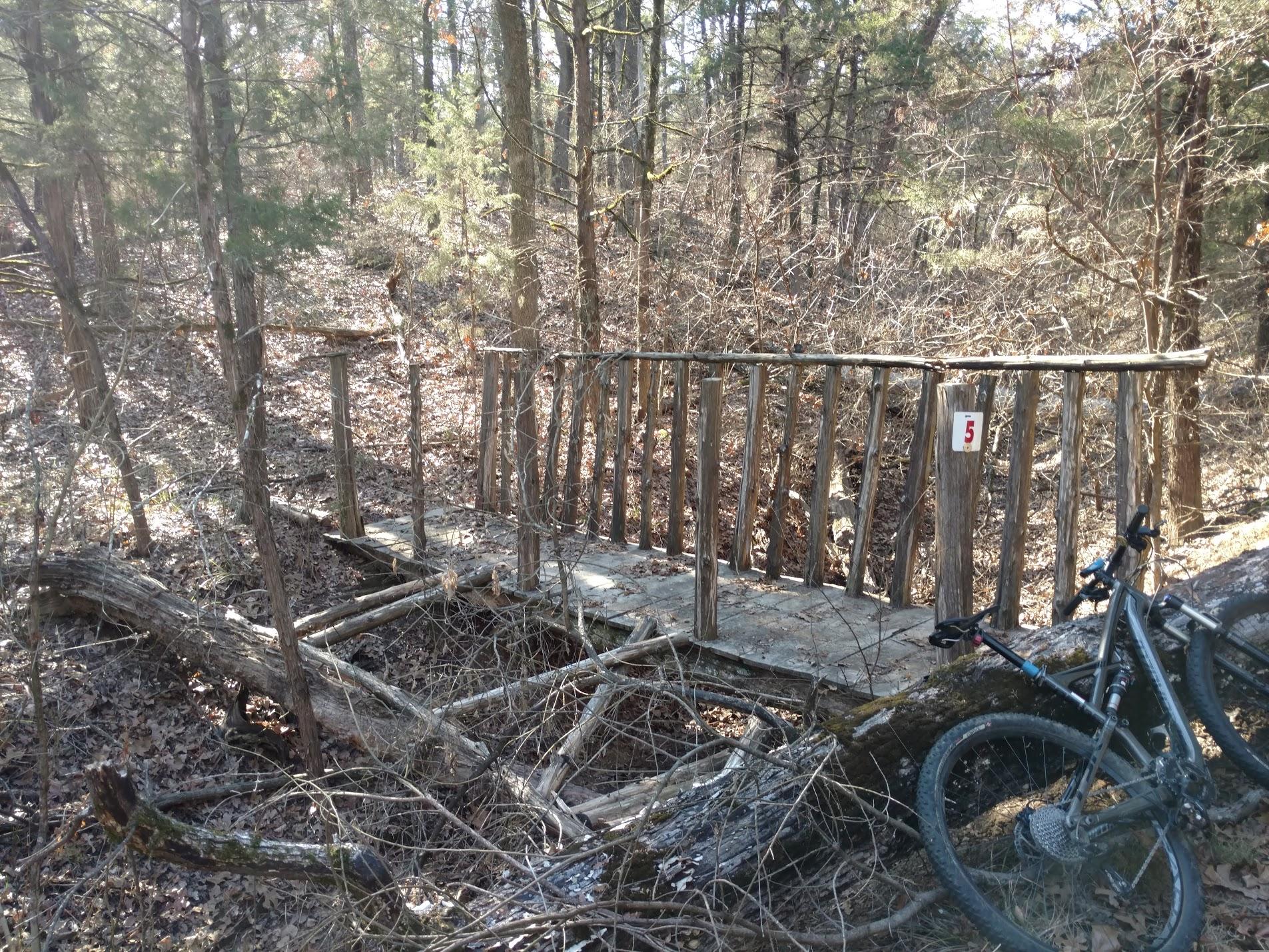 A rustic wooden bridge in a forest setting, surrounded by trees and underbrush. A mountain bike leans against a fallen log nearby. The bridge features a simple design with wooden railings and has a numbered marker labeled "5." The landscape is covered in fallen leaves, indicating an autumn or early winter season. Lake Fannin Mountain Bike Trail mountain bike trail.
