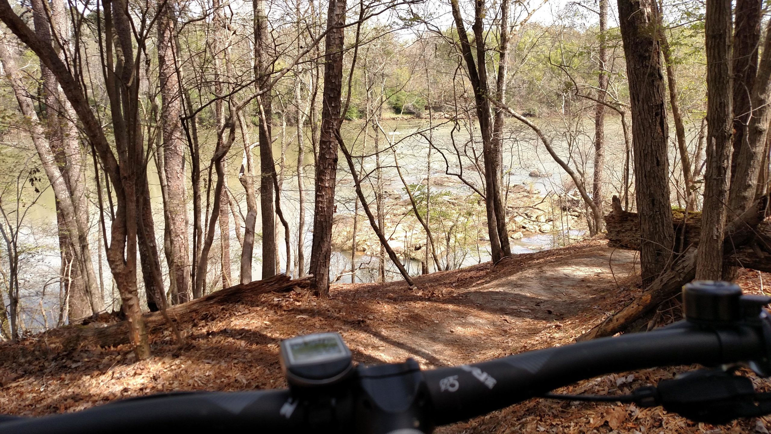 A scenic view of a river surrounded by trees, captured from the perspective of a bicycle handlebar. The landscape features a winding path leading towards the water, with rocky outcrops visible in the river. The ground is covered with fallen leaves, and the atmosphere conveys a peaceful, natural setting. Harbison State Forest mountain bike trail.