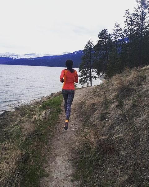 A person running on a dirt path beside a lake, surrounded by grassy areas and trees. Snow-capped mountains are visible in the background under a cloudy sky. The runner is wearing a bright orange top and patterned leggings. Evely Park Trail mountain bike trail.
