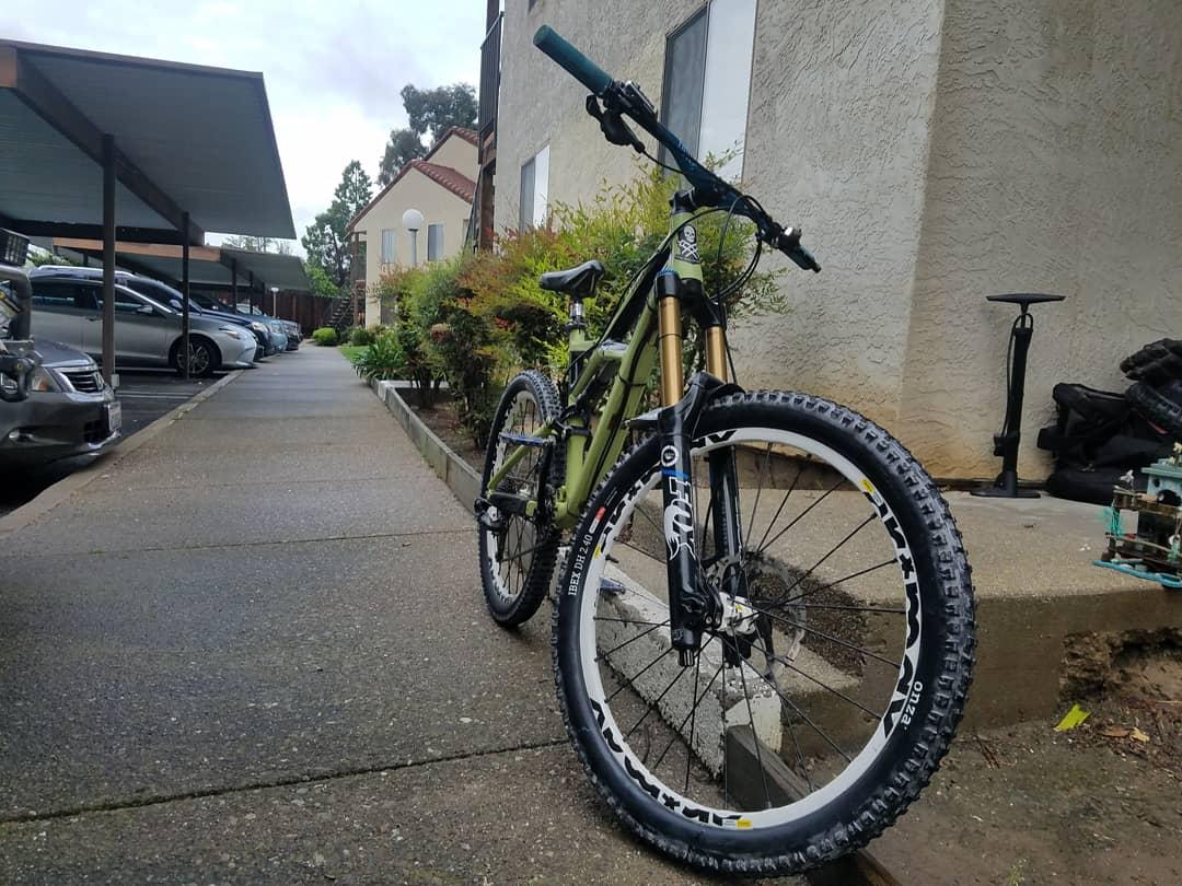 Specialized Enduro: A green mountain bike parked on a sidewalk near an apartment complex. The bike features a front suspension fork and wide tires with a patterned tread. In the background, there are parked cars and shrubs lining the walkway.