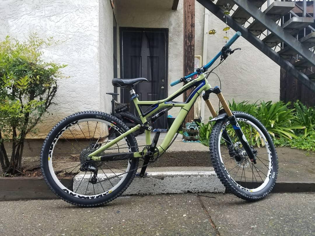 Specialized Enduro: A green mountain bike with front suspension parked on a pavement outside a building. The bike features black tires with white rims and colorful handlebars. In the background, there is a staircase and some greenery.