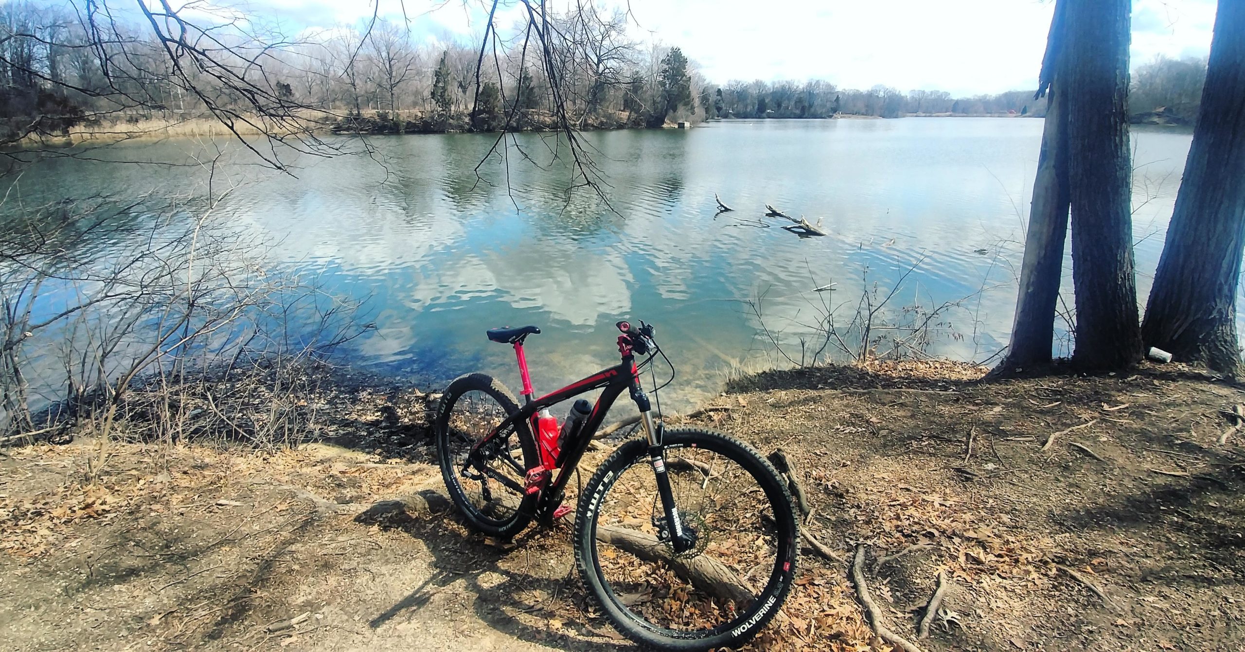 A mountain bike rests on the shore next to a calm lake surrounded by trees and bare branches, with reflections of clouds on the water's surface. Hines Park Trail mountain bike trail.