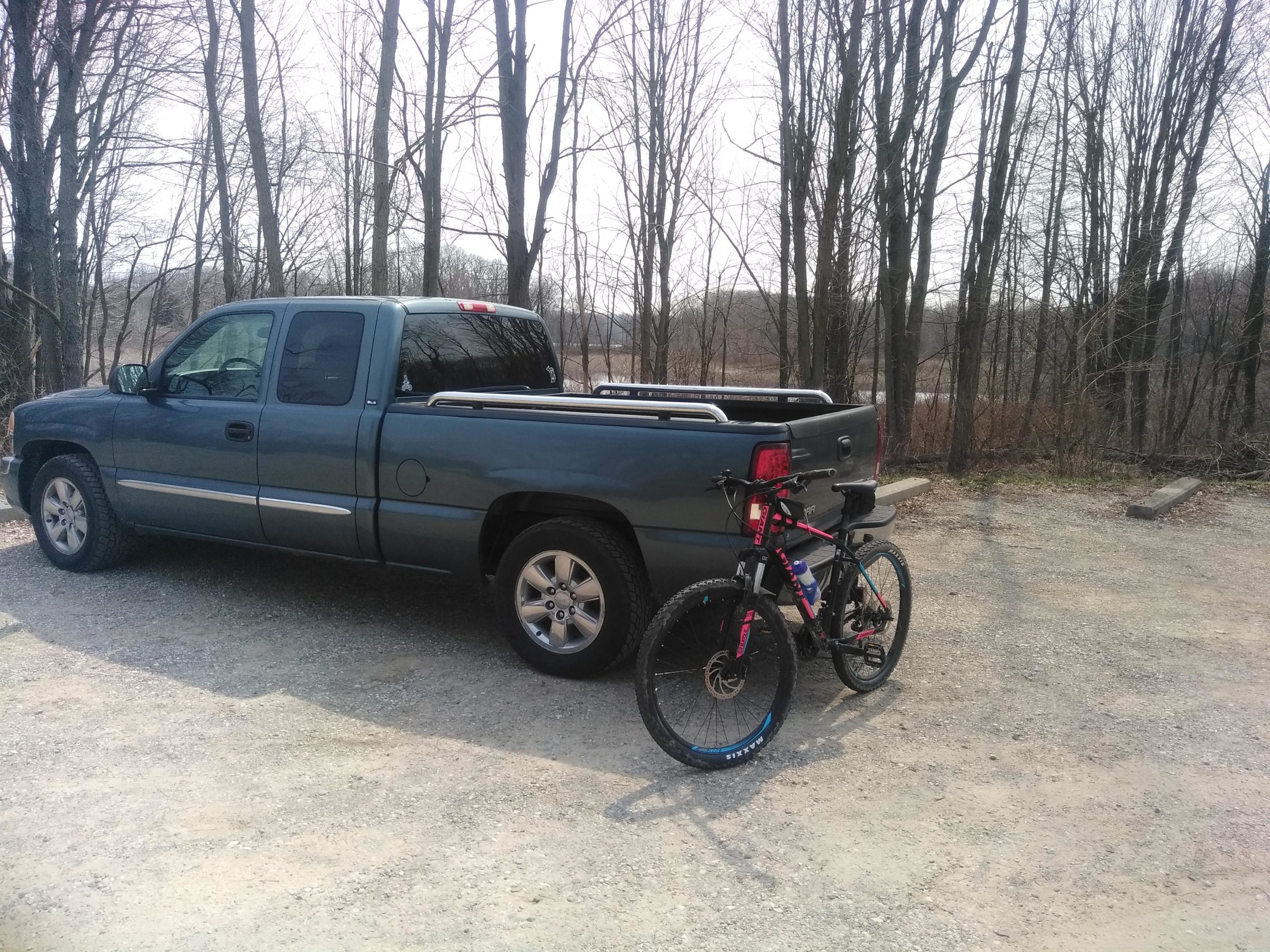 A blue pickup truck parked on a gravel surface, with a mountain bike leaning against its rear. Leafless trees are visible in the background, hinting at an early spring setting. DTE Energy Foundation Trail mountain bike trail.