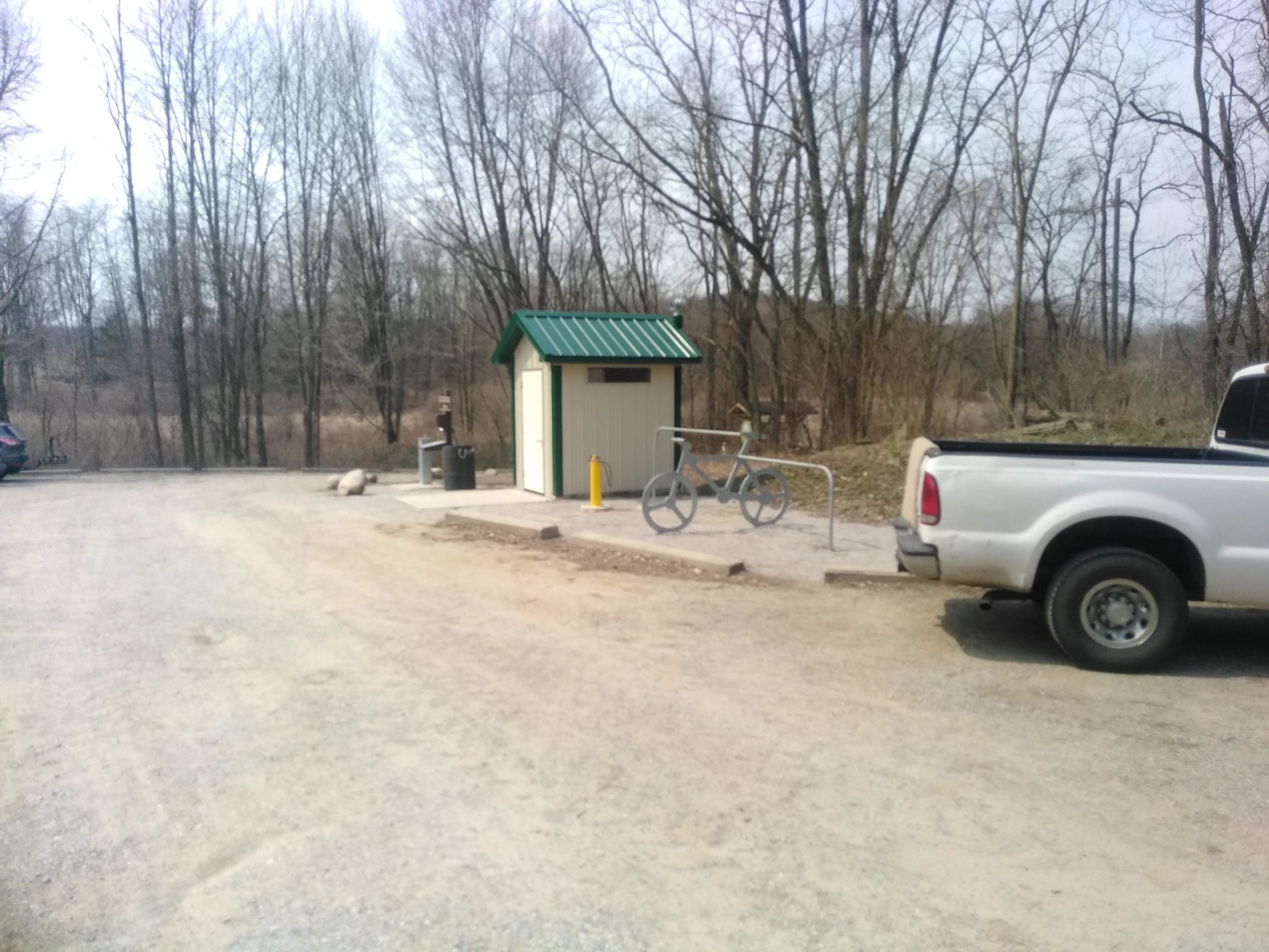 A gravel parking area with a white truck parked to the right and a small storage building with a green roof on the left. Nearby, there is a bike rack shaped like a bicycle and a trash receptacle. The background features leafless trees, indicating early spring or late winter. DTE Energy Foundation Trail mountain bike trail.