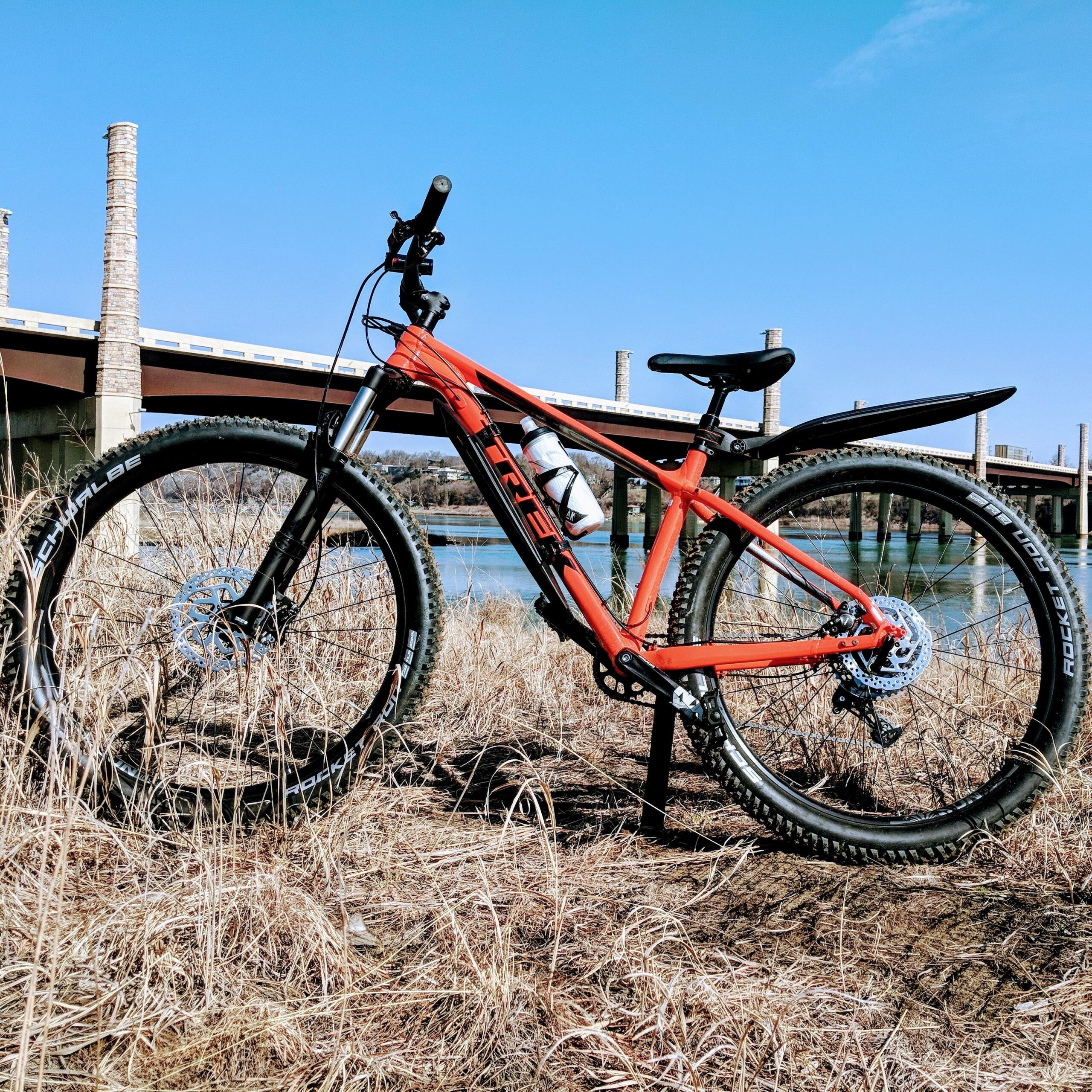 A bright orange mountain bike is leaning on a grassy area near a body of water, with a bridge visible in the background under a clear blue sky. The bike features thick tires and a water bottle mounted on the frame.