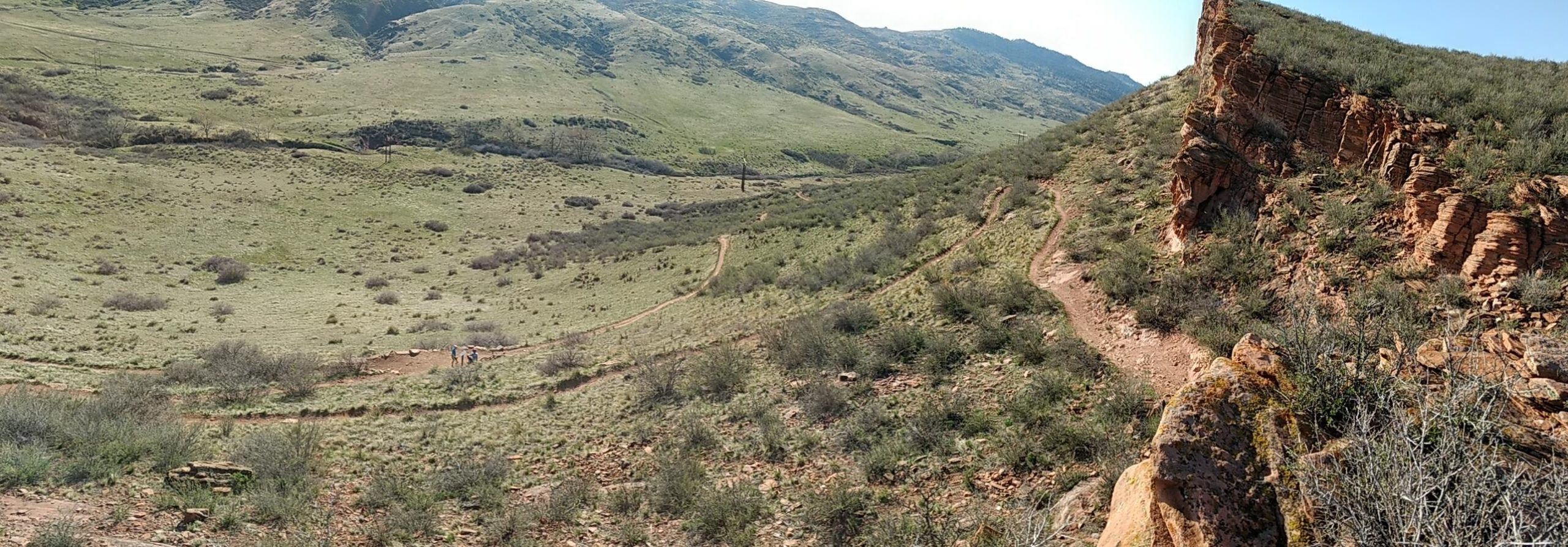 A panoramic view of a green landscape with rolling hills and rocky formations. A winding dirt trail leads through the grassy area, and several small shrubs and plants are scattered throughout the scene. In the distance, a few hikers can be seen on the path, enjoying the natural surroundings under a clear blue sky. Blue Sky mountain bike trail.