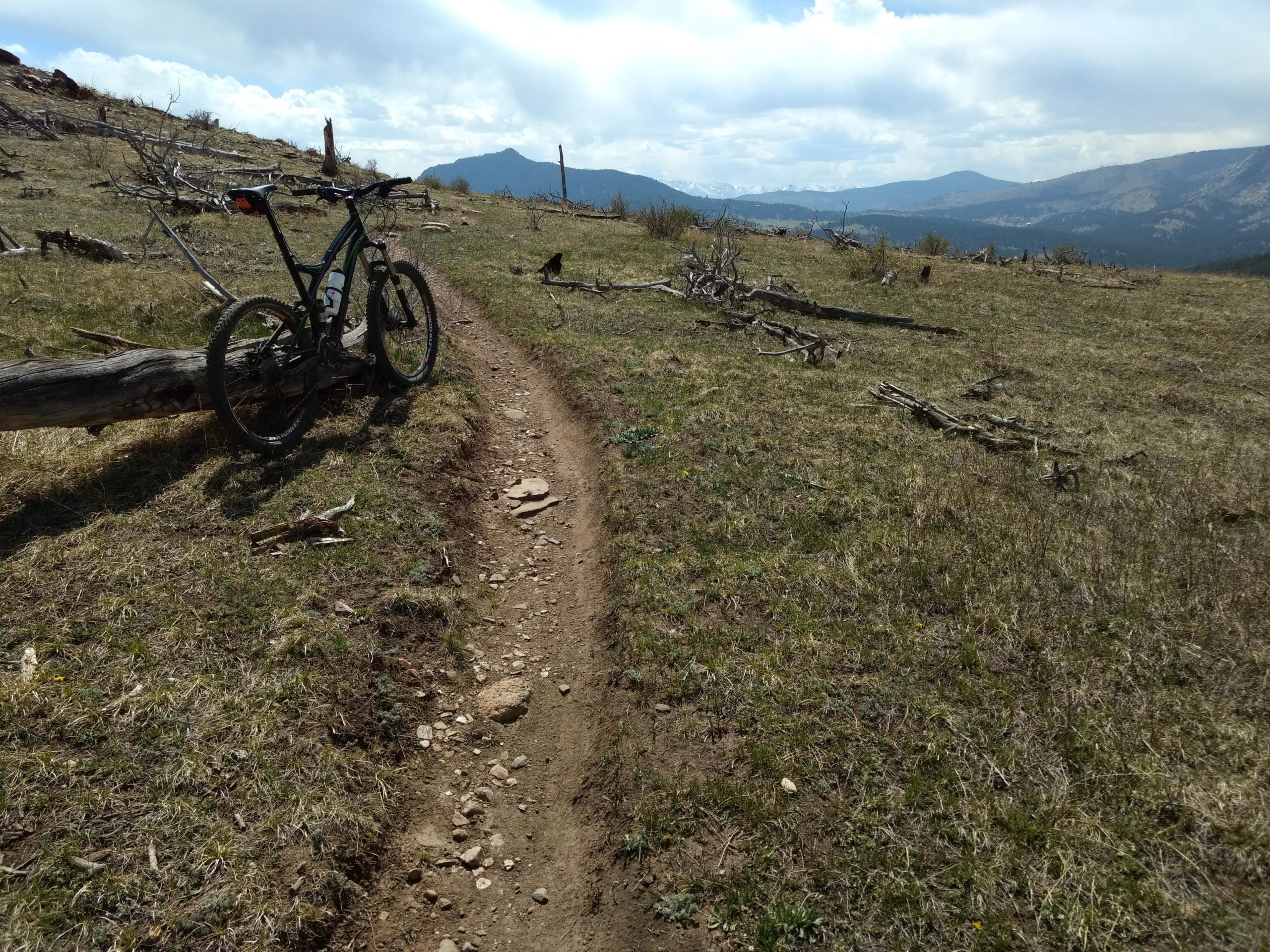 A mountain bike is parked beside a narrow dirt trail that winds through a grassy area with scattered fallen branches. In the background, rolling hills and distant mountains are visible under a partly cloudy sky. Ginny Trail mountain bike trail.