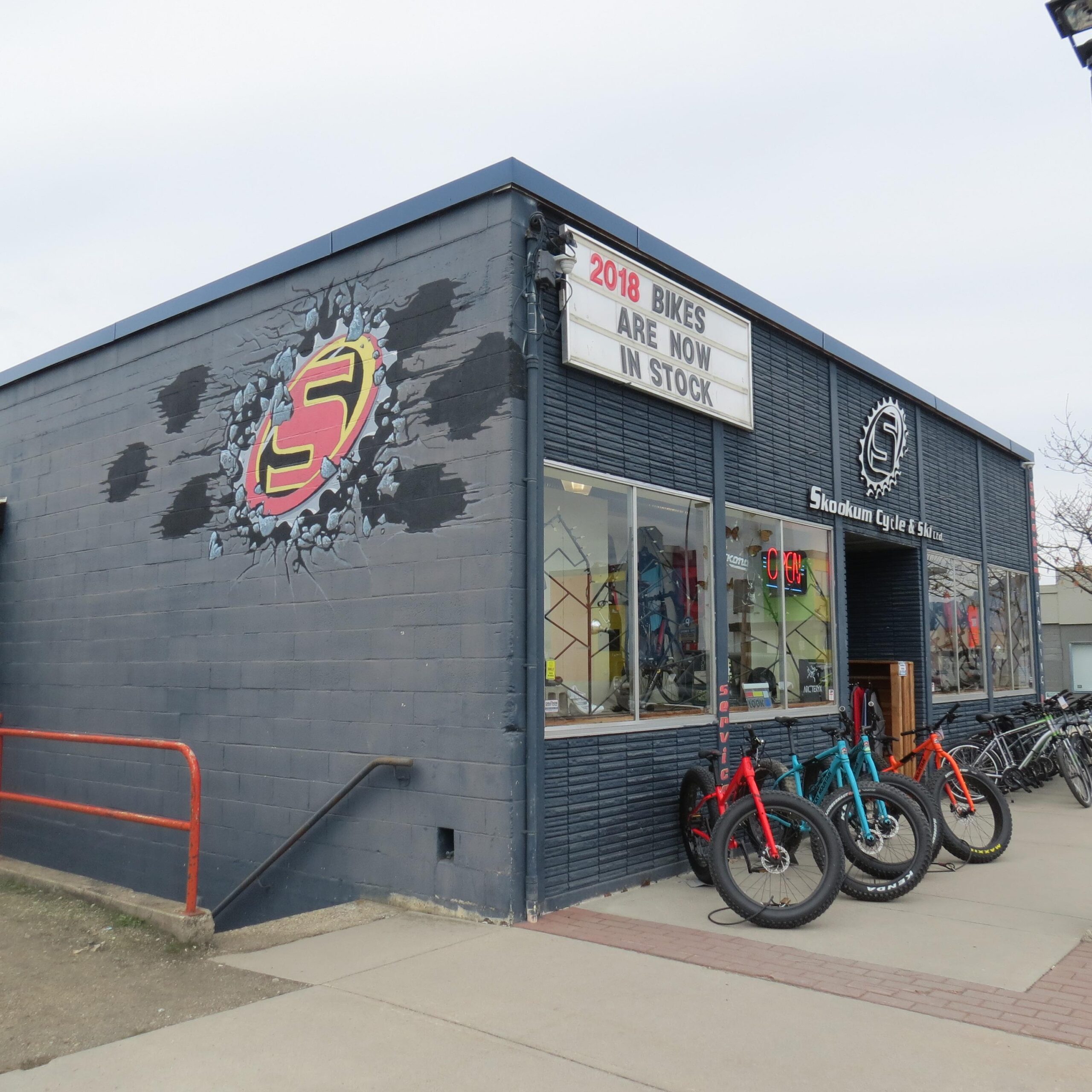 Exterior view of a bicycle shop named "Skookum Cycle & Ski," featuring a colorful logo painted on the wall. A sign above announces "2018 Bikes Are Now In Stock." In front of the store, several bicycles, including fat bikes in red and blue, are displayed on the sidewalk. The building has a modern look with large windows, allowing visibility of bike models inside.