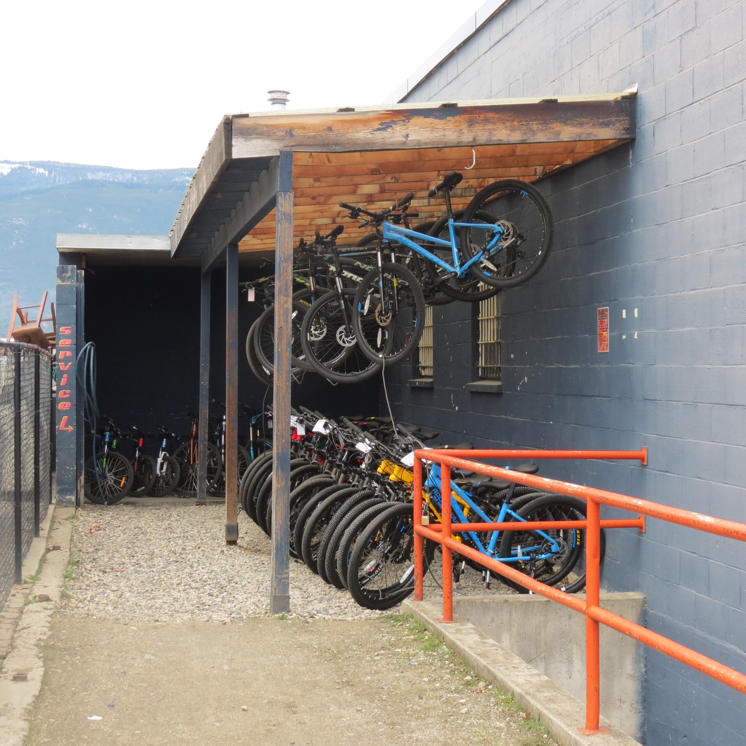 A covered bike storage area against a blue wall, featuring several bicycles hanging from the roof and others stored on the ground. An orange railing runs along the pathway, with gravel and a grassy edge visible. The background shows a mountainous landscape.