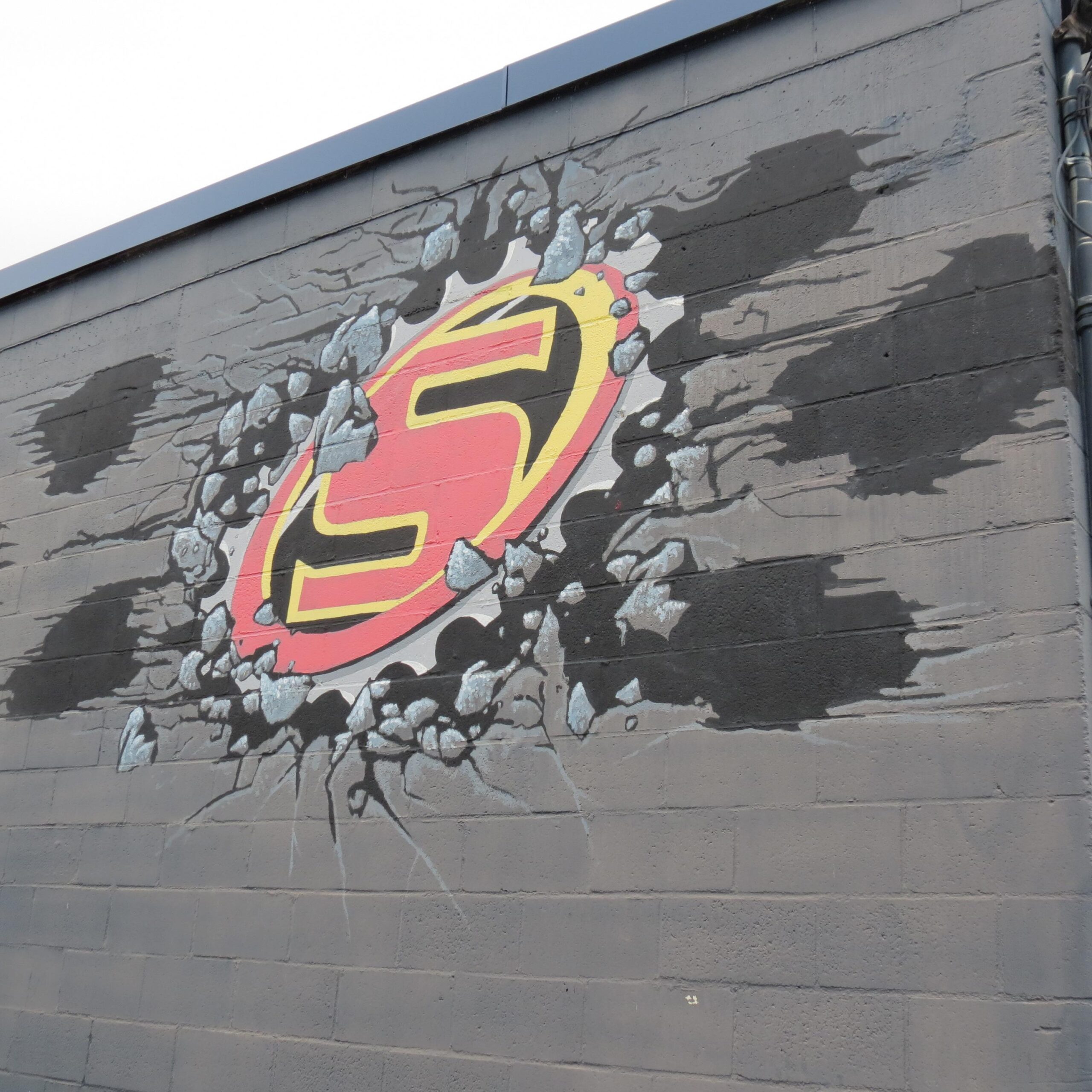 A colorful mural on a gray wall featuring a superhero emblem breaking through the surface, surrounded by cracks and debris. The emblem is circular with a red background and a black stylized letter "S" at its center, evoking a dynamic and bold aesthetic.