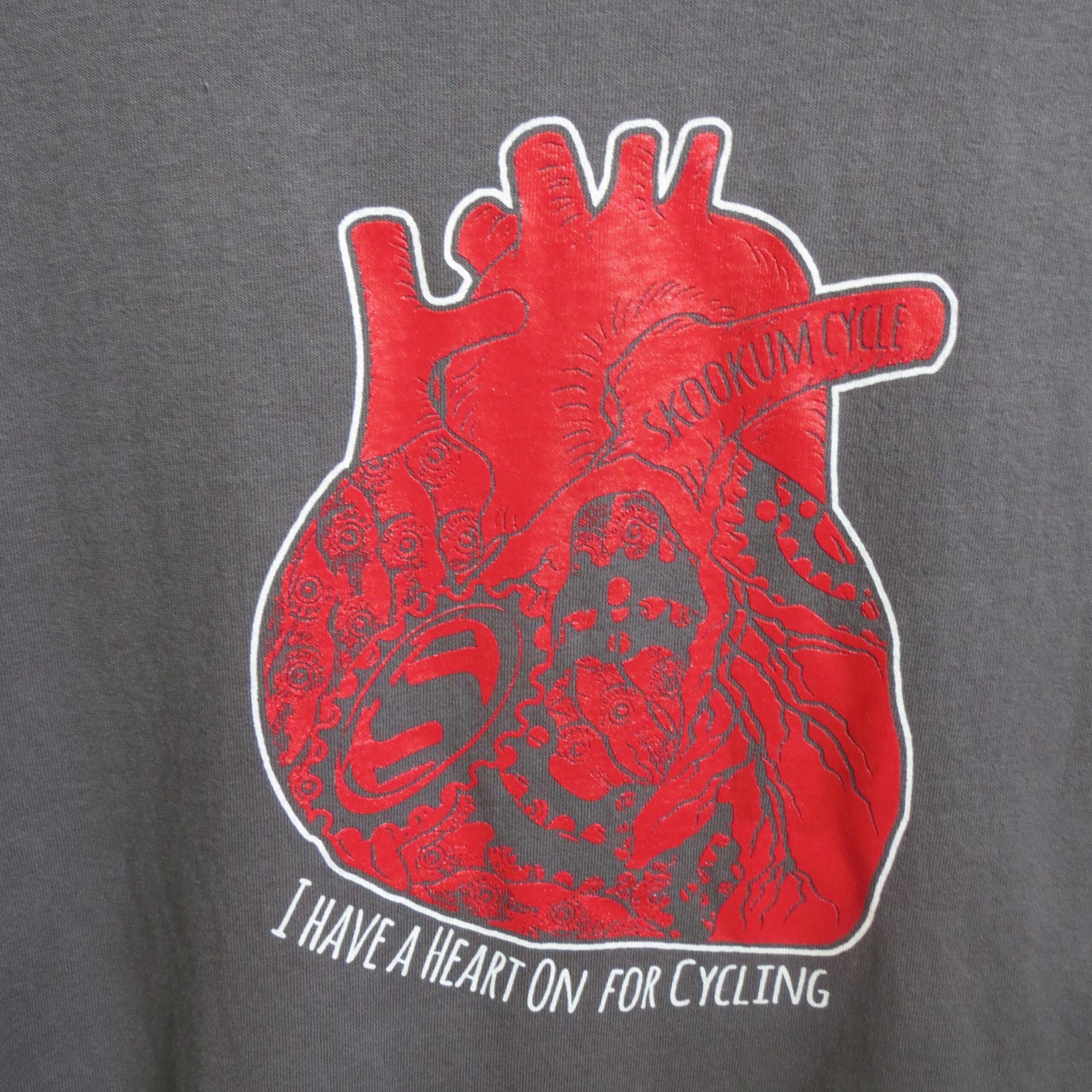 A graphic design featuring a stylized heart in red, with intricate patterns and cycling-related imagery, set against a gray background. The text reads, "I HAVE A HEART ON FOR CYCLING," along with the words "SKOOKUM CYCLE" integrated into the design.